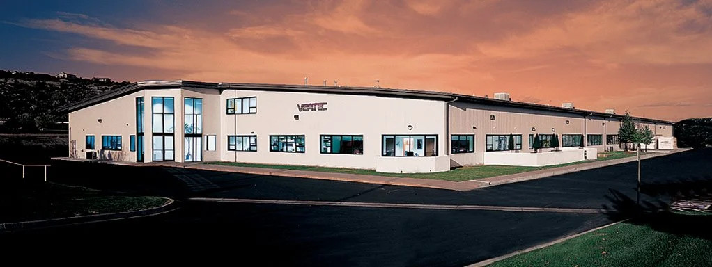 Large industrial or office building with a white facade, multiple windows, and a corner entrance, under an orange sunset sky.
