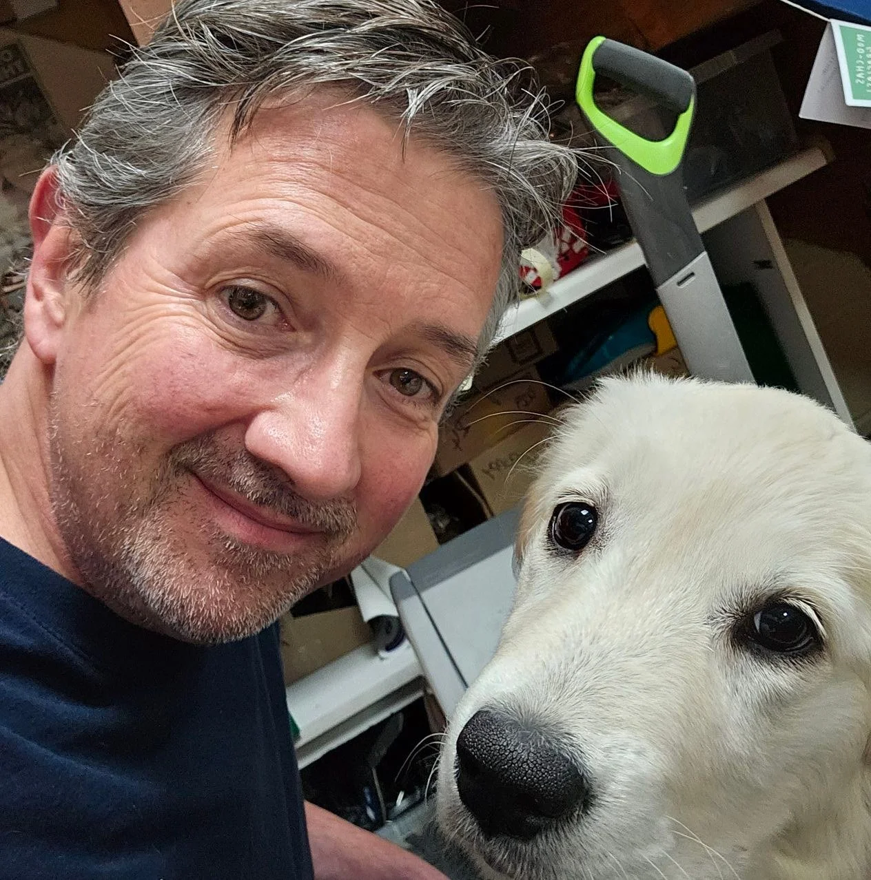 A man with gray hair and a beard taking a selfie with a white dog, possibly a retriever, indoors with shelves and various items in the background.