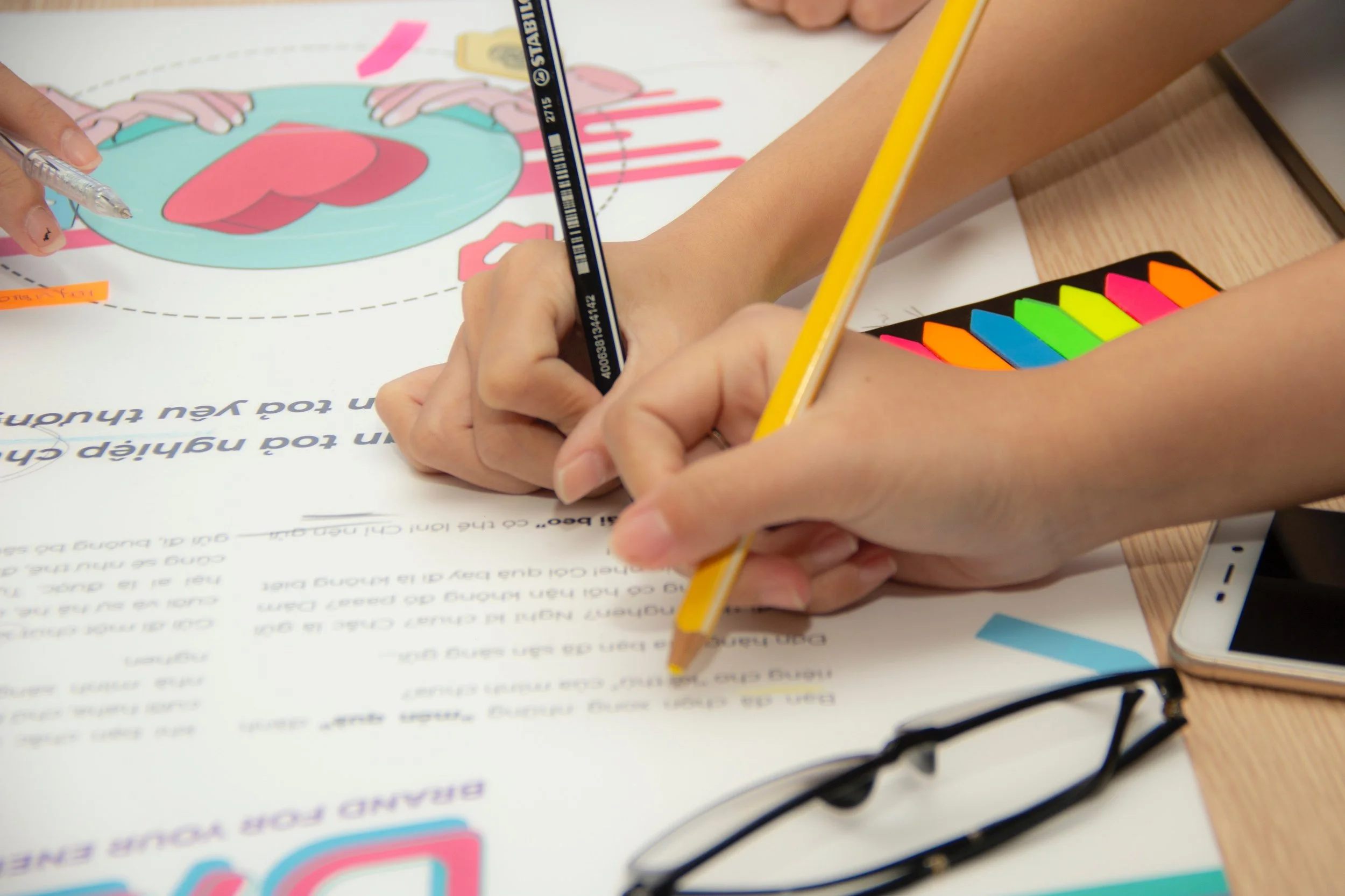 A person holding a yellow pencil and a silver pen, working on a colorful educational poster with diagrams of the human body, including a heart and digestive system, on a desk.