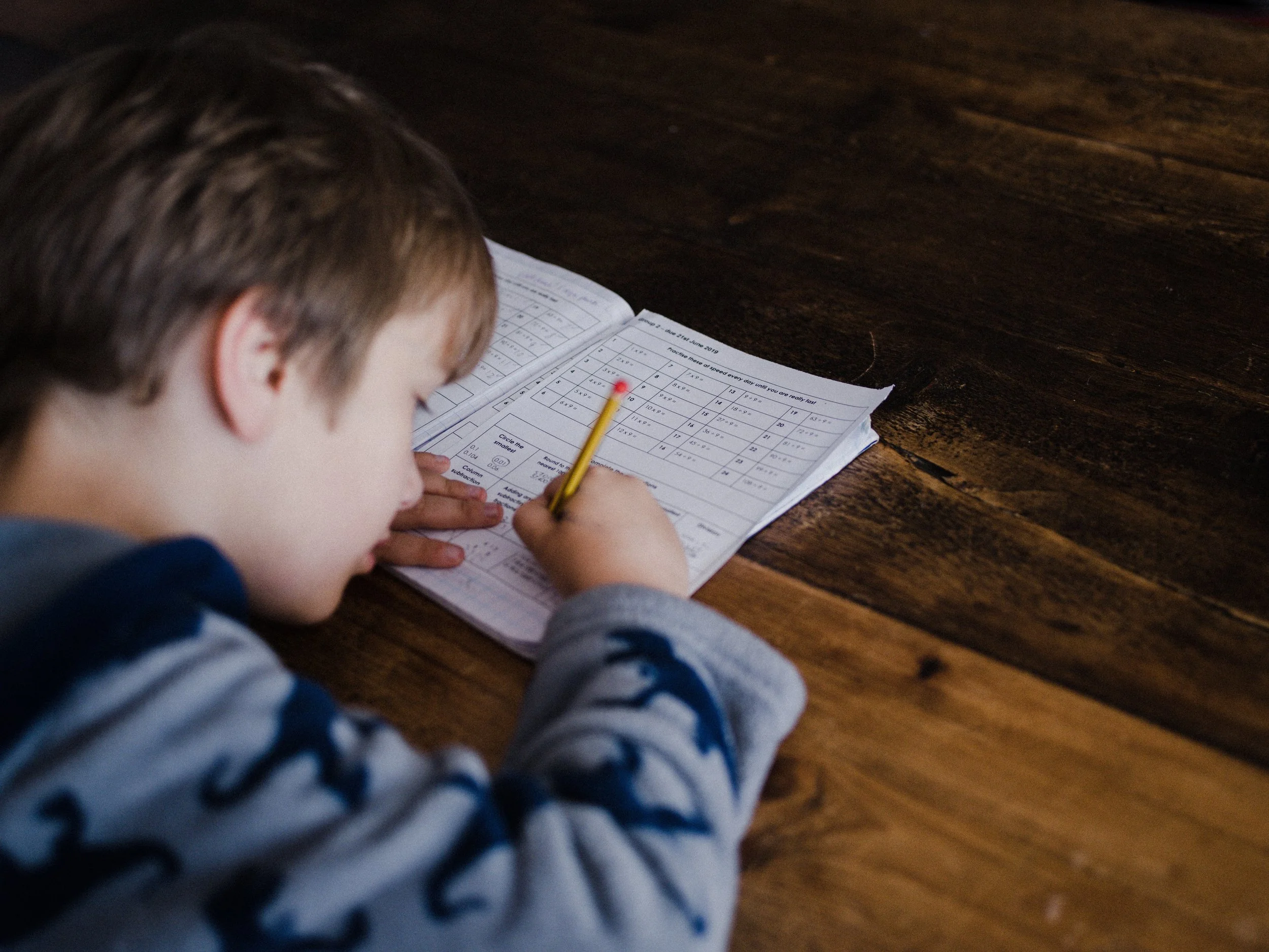 A young boy with short brown hair, seen from the side, is writing or drawing on a worksheet with a yellow pencil. The worksheet is open on a wooden table.