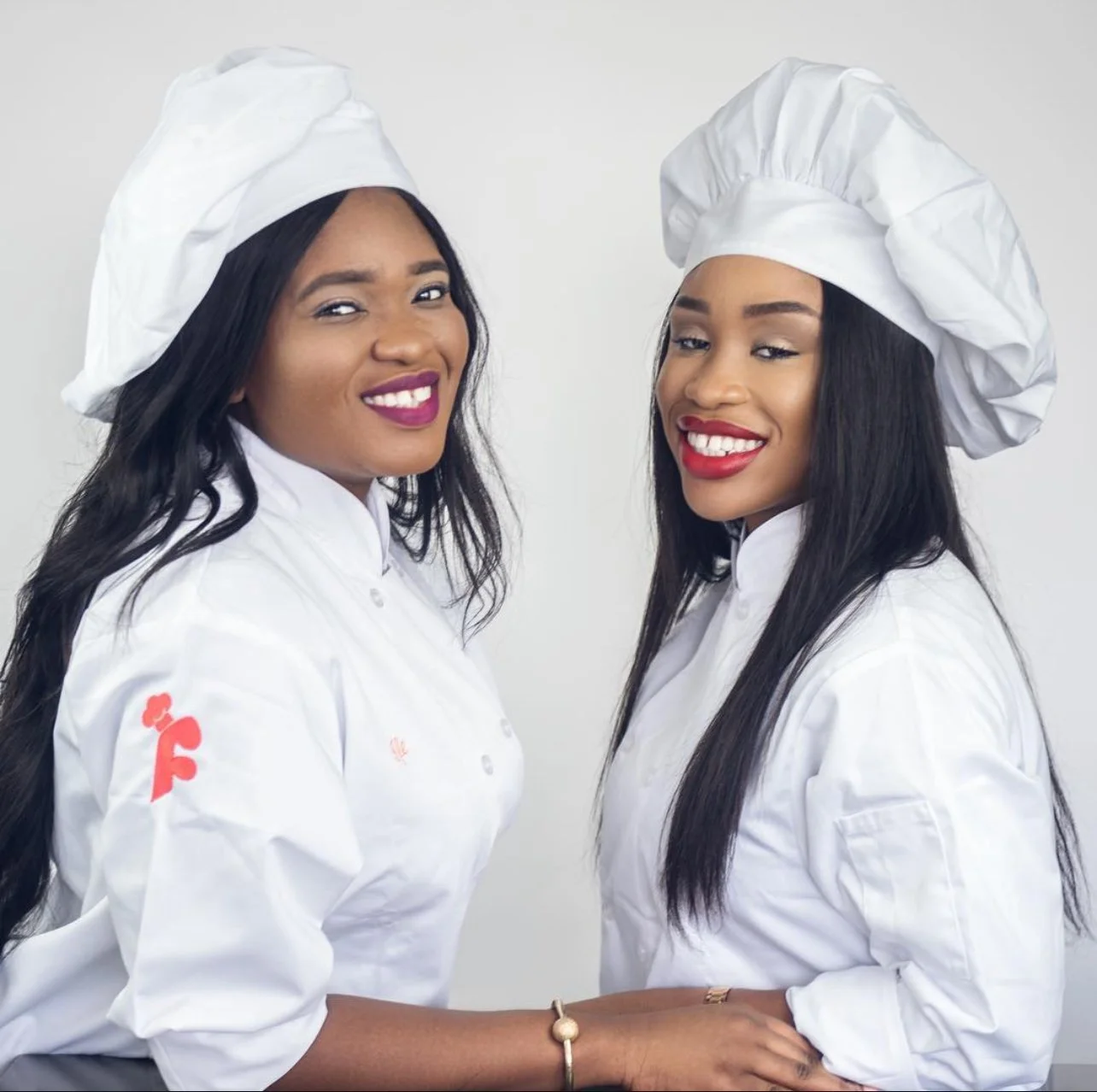 Two women dressed as chefs in white uniforms and chef hats, smiling and standing close together.