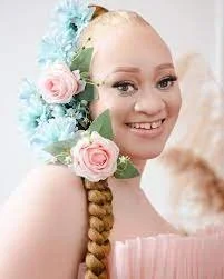 Young woman smiling with a floral headpiece of pink roses and blue flowers, styled in long braided hair, wearing a pink top.