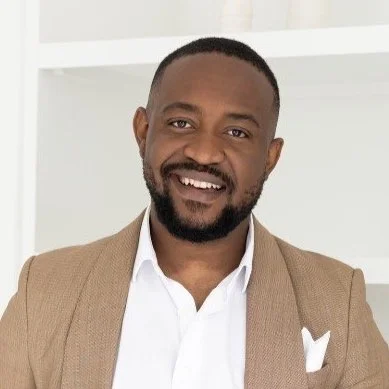 A smiling Black man with a short beard, wearing a beige blazer and white shirt, standing indoors in front of a white background.