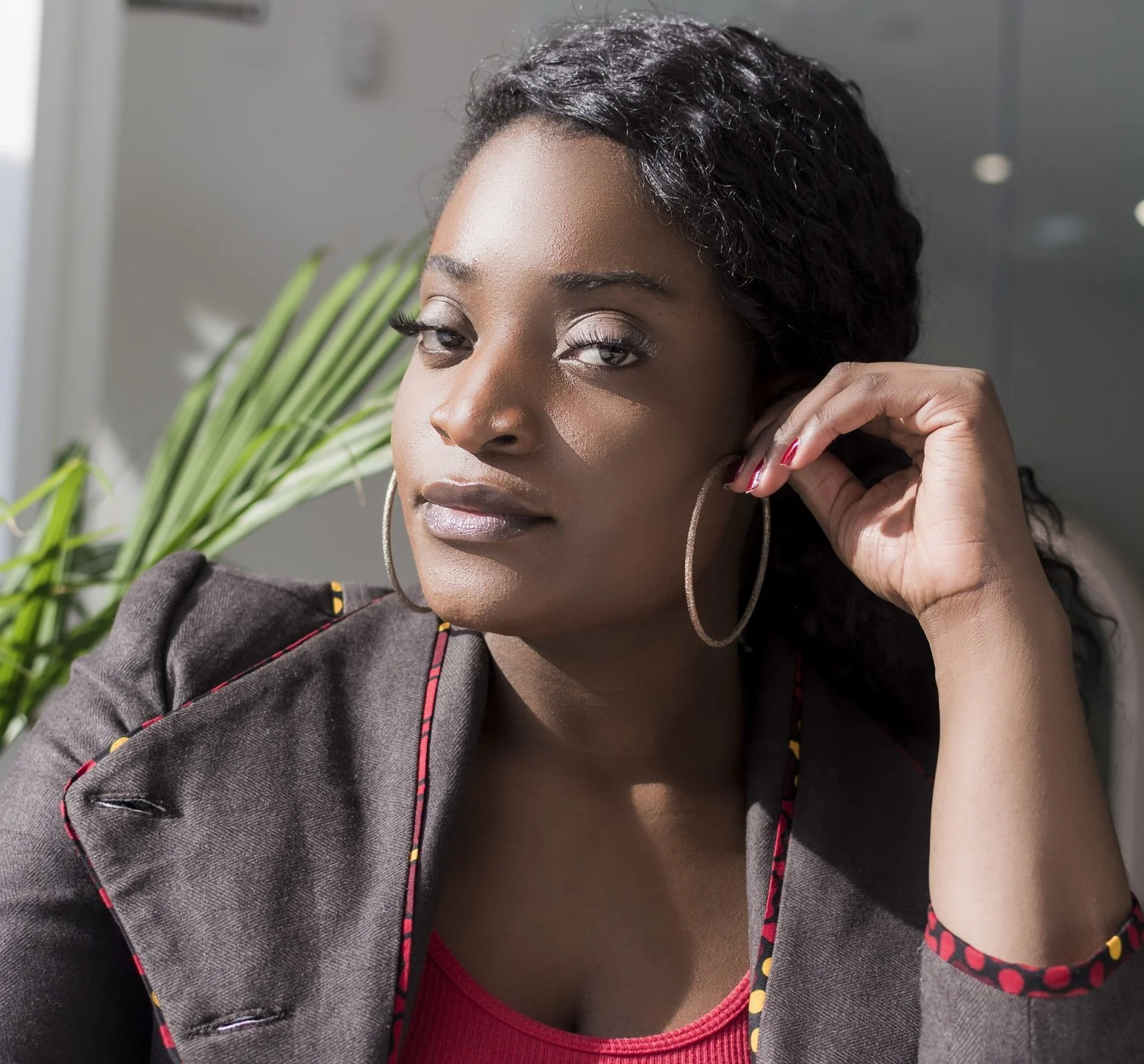 A woman with dark curly hair, wearing large hoop earrings, a dark blazer with colorful trim, and a red top, looking confidently at the camera while adjusting her earring.