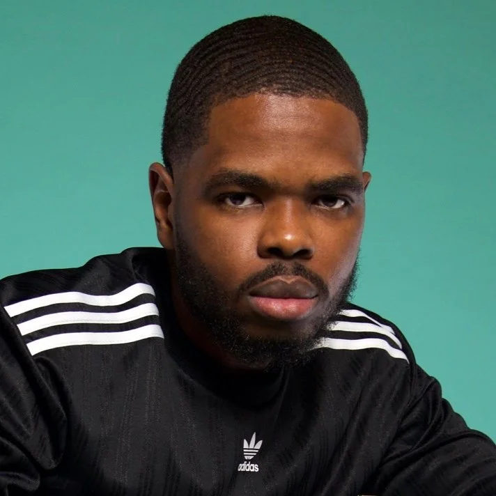 A young man with dark skin, short curly hair, and a beard looking at the camera. He is wearing a black Adidas sports shirt with white stripes on the shoulders, against a solid green background.