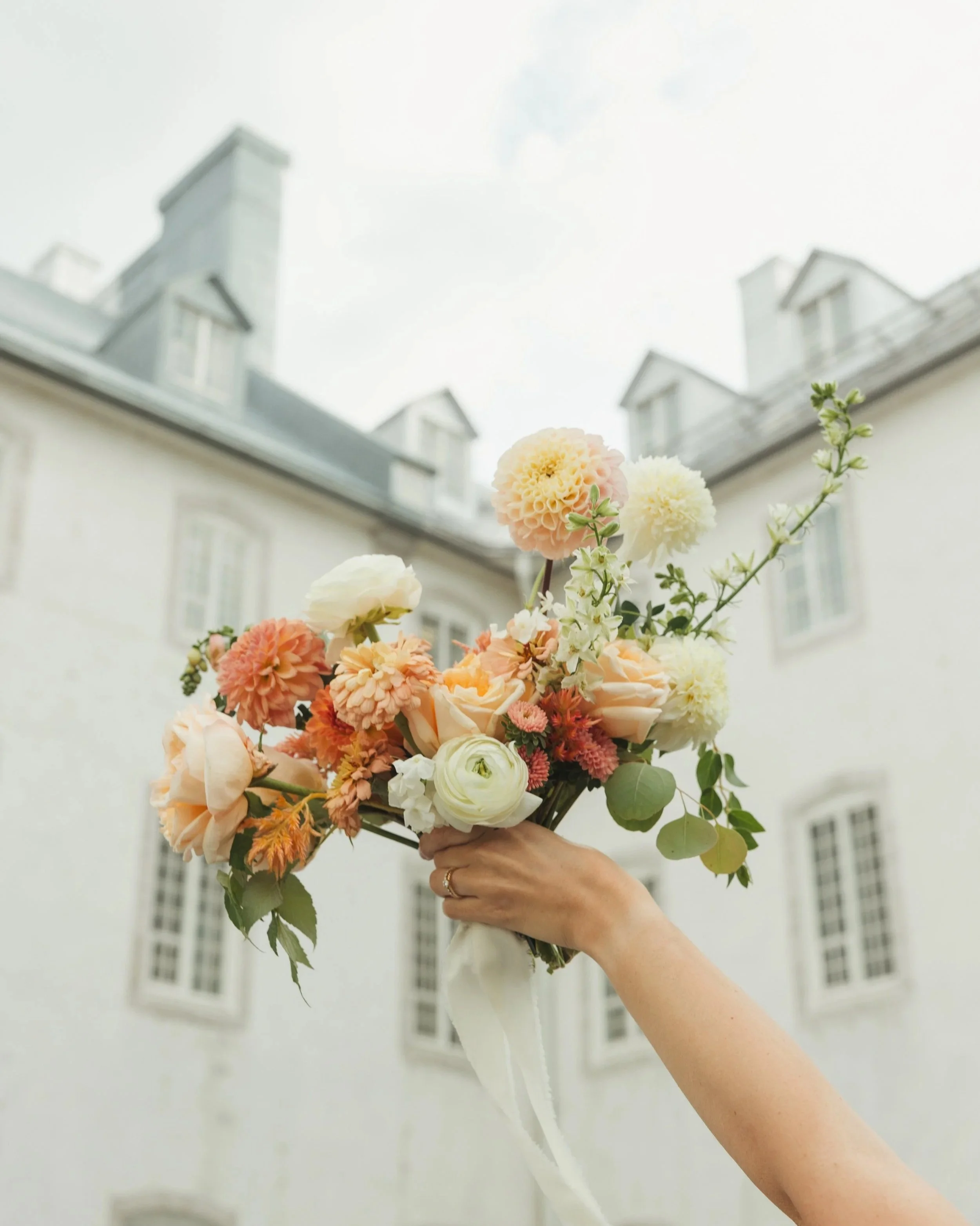 Main tenant un bouquet de fleurs de différentes couleurs, tenu devant un bâtiment blanc avec des fenêtres blanches, sous un ciel clair.