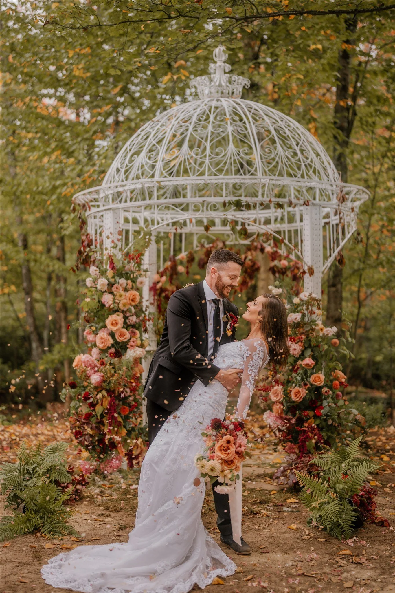Un couple de mariés danse sous une structure en fer blanc ornée de fleurs, en pleine forêt lors d'un mariage automnal.