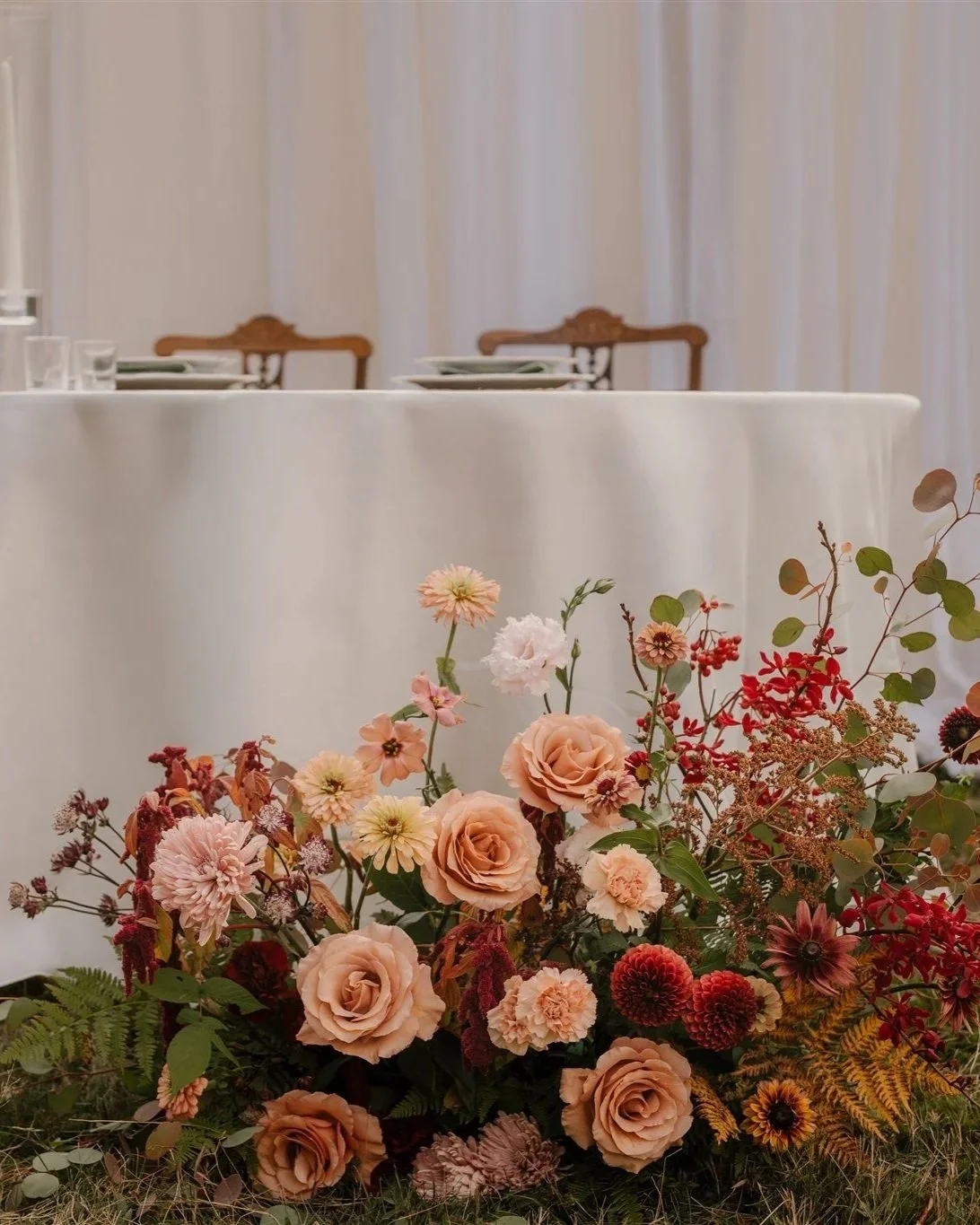 Arrangement floral composé de roses crème, dahlias, cosmos, et feuillage varié, placé devant une table habillée d'une nappe blanche et entourée de chaises en bois.