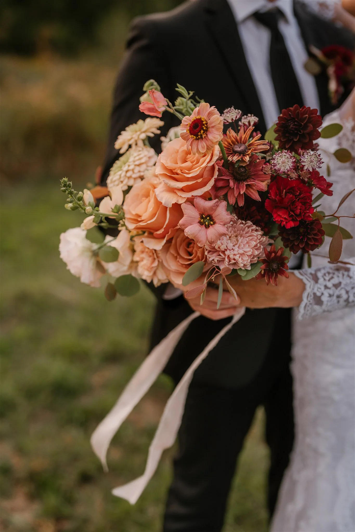Une personne en robe de mariée tenant un bouquet de fleurs pastel et rouges, avec un homme en costume noir en arrière-plan.