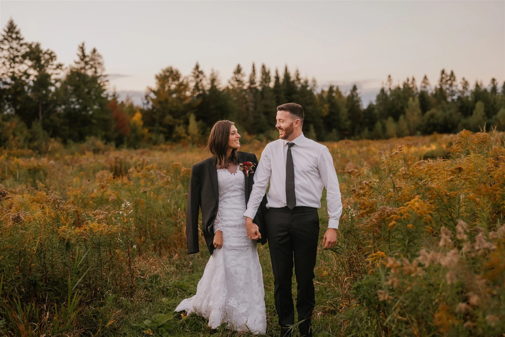 Un couple marchant main dans un champ de fleurs sauvages lors d'un coucher de soleil, la femme en robe de mariée et le homme en chemise blanche et cravate.