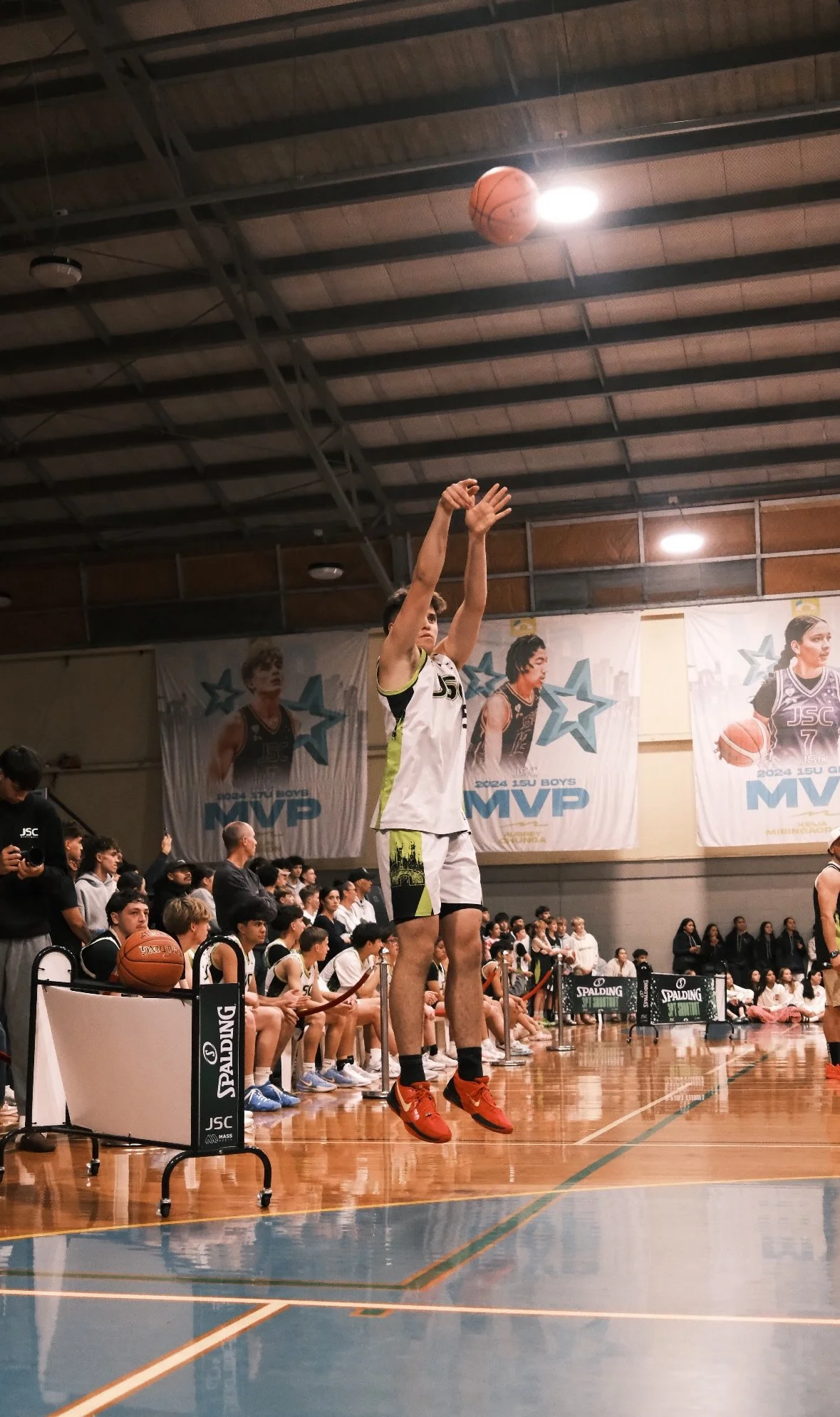 Young basketball player in a white and black uniform taking a jump shot on an indoor basketball court, surrounded by seated teammates and spectators, with banners and posters on the wall.