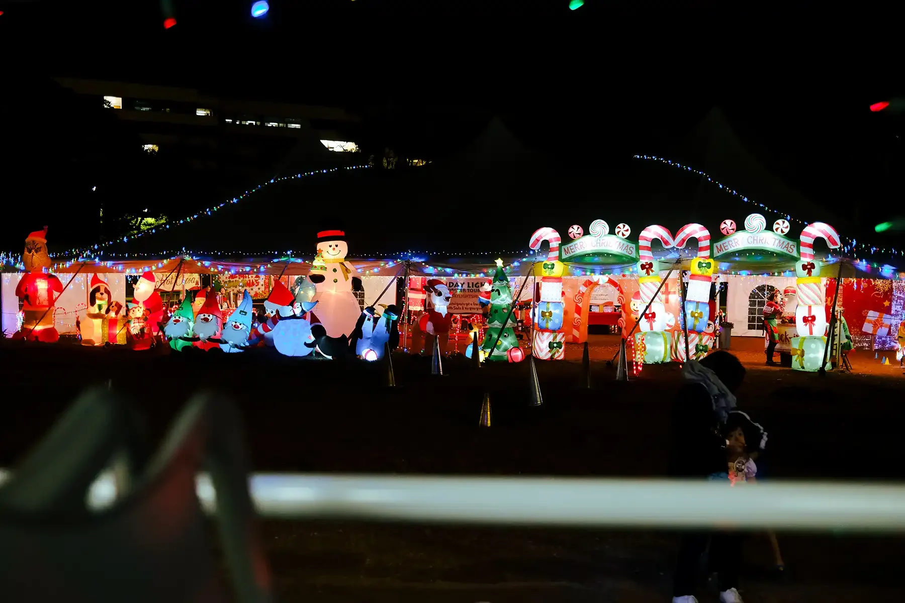 Colorful Christmas light display featuring inflatable decorations including Santa Claus, snowmen, elves, and Christmas trees, with a tent and people in the background at night.