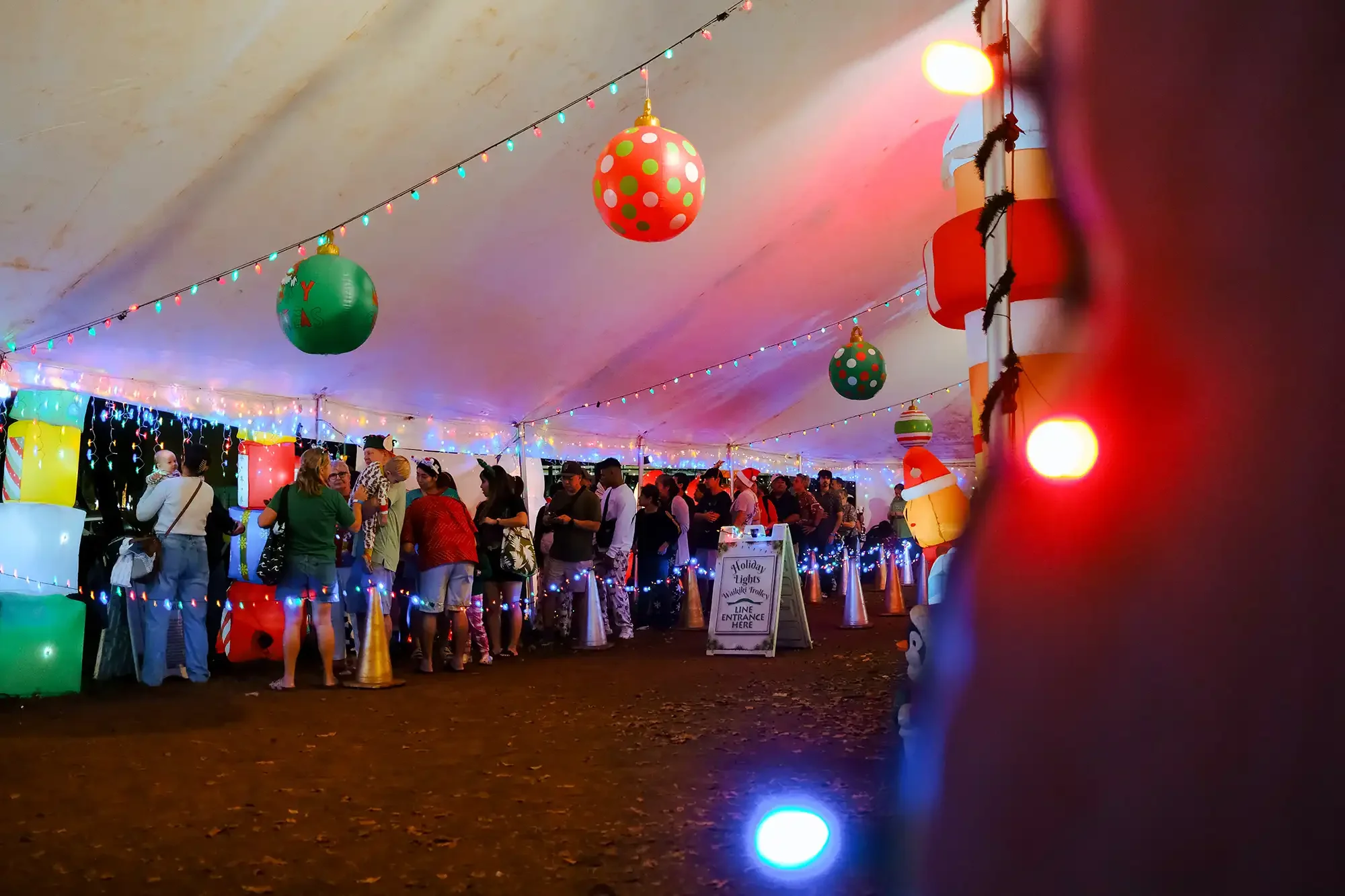Christmas-themed party under a decorated tent with colorful hanging ornaments, string lights, and a line of people waiting at a festive distribution table.