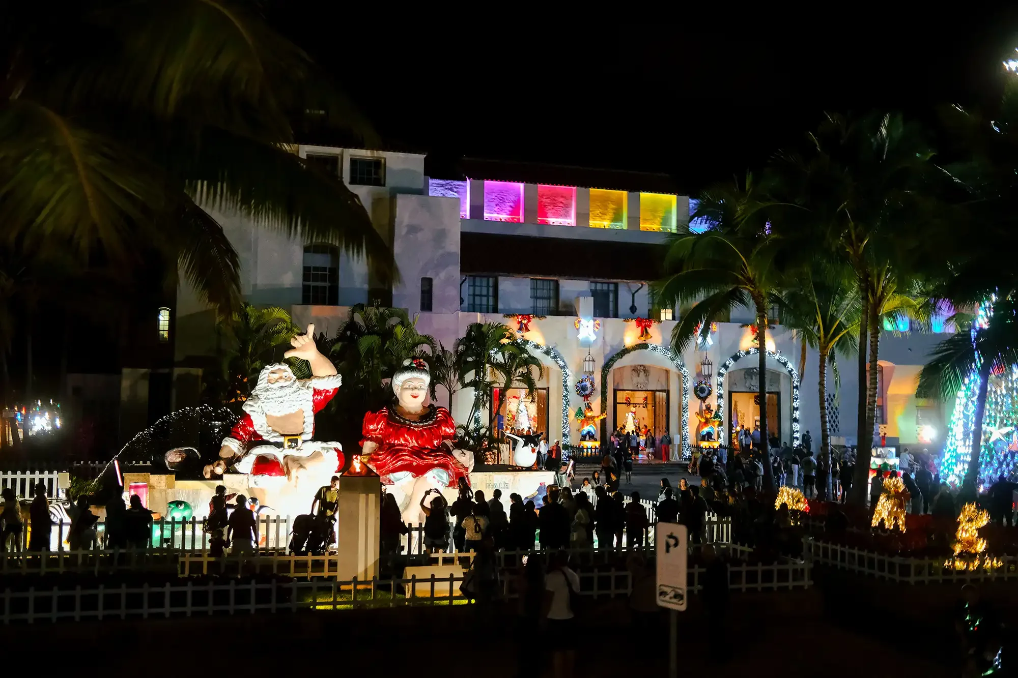 At the Honolulu Hale, Nighttime scene of a building decorated with colorful Christmas lights, featuring large Santa Claus and Mrs. Claus statues, illuminated arches, and a crowd of people in front.