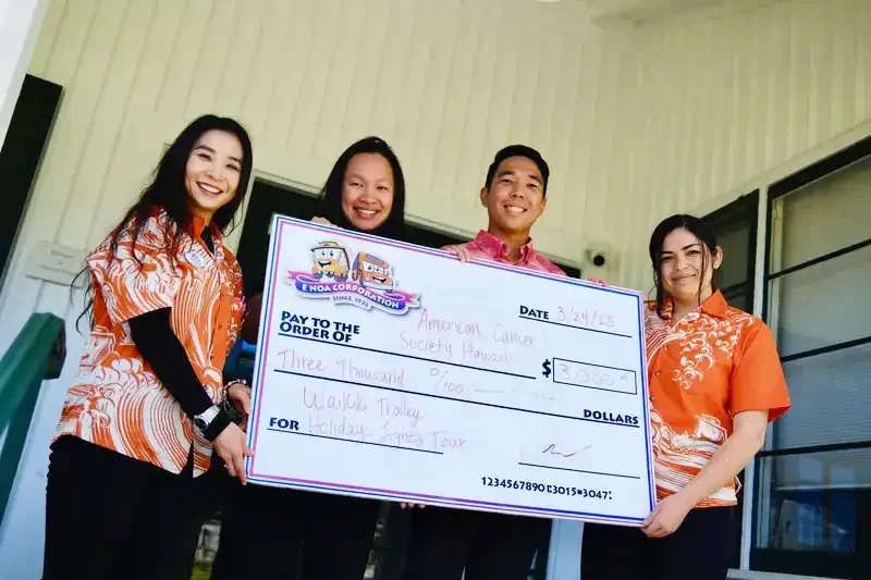 Four people, three women and one man, standing outdoors and holding a large check made out to the American Cancer Society Society Hawaiian for $3,000, dated April 3, 2023, during daytime.
