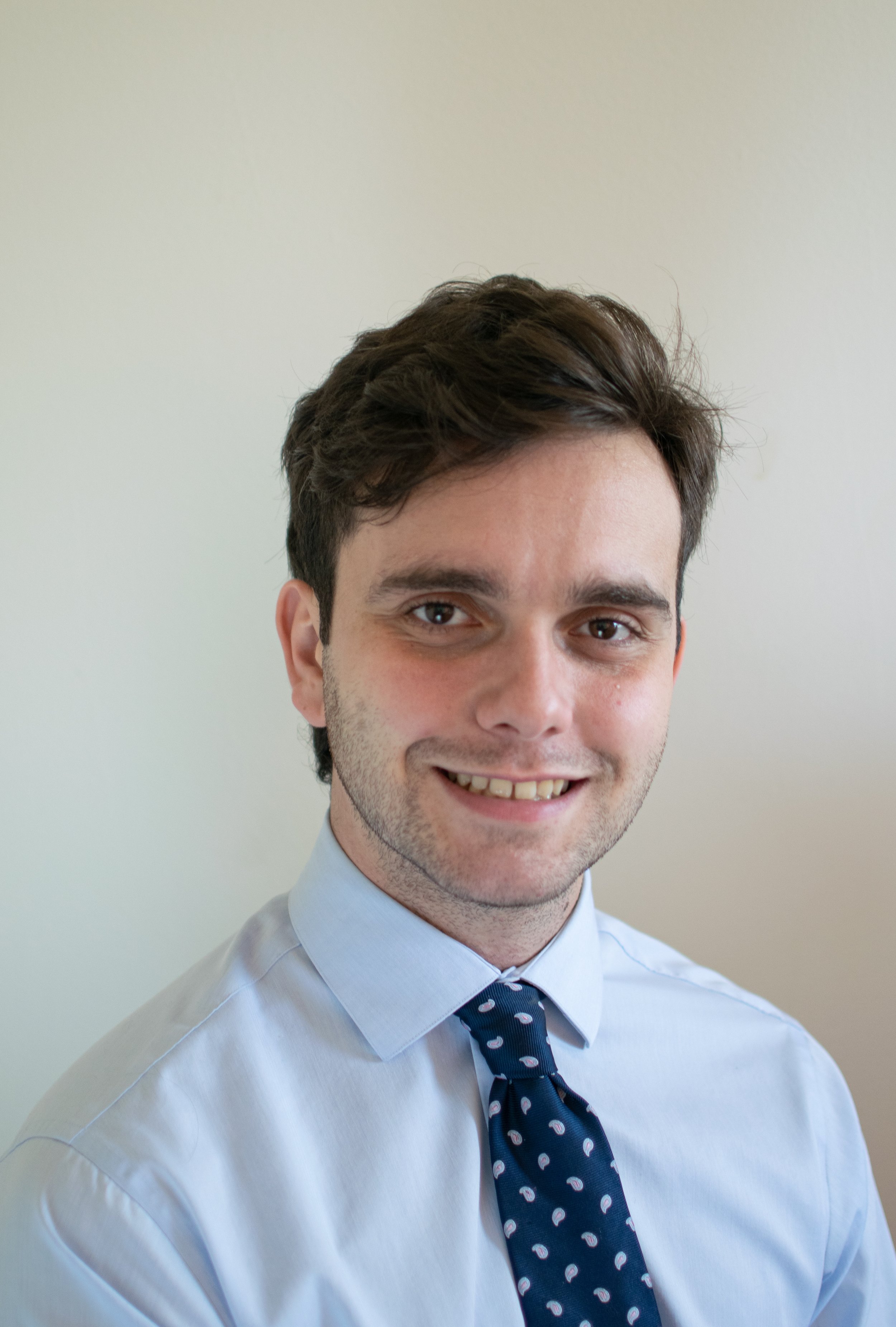 A young man with dark brown hair, wearing a light blue dress shirt and a navy blue polka dot tie, smiling at the camera against a plain light background.