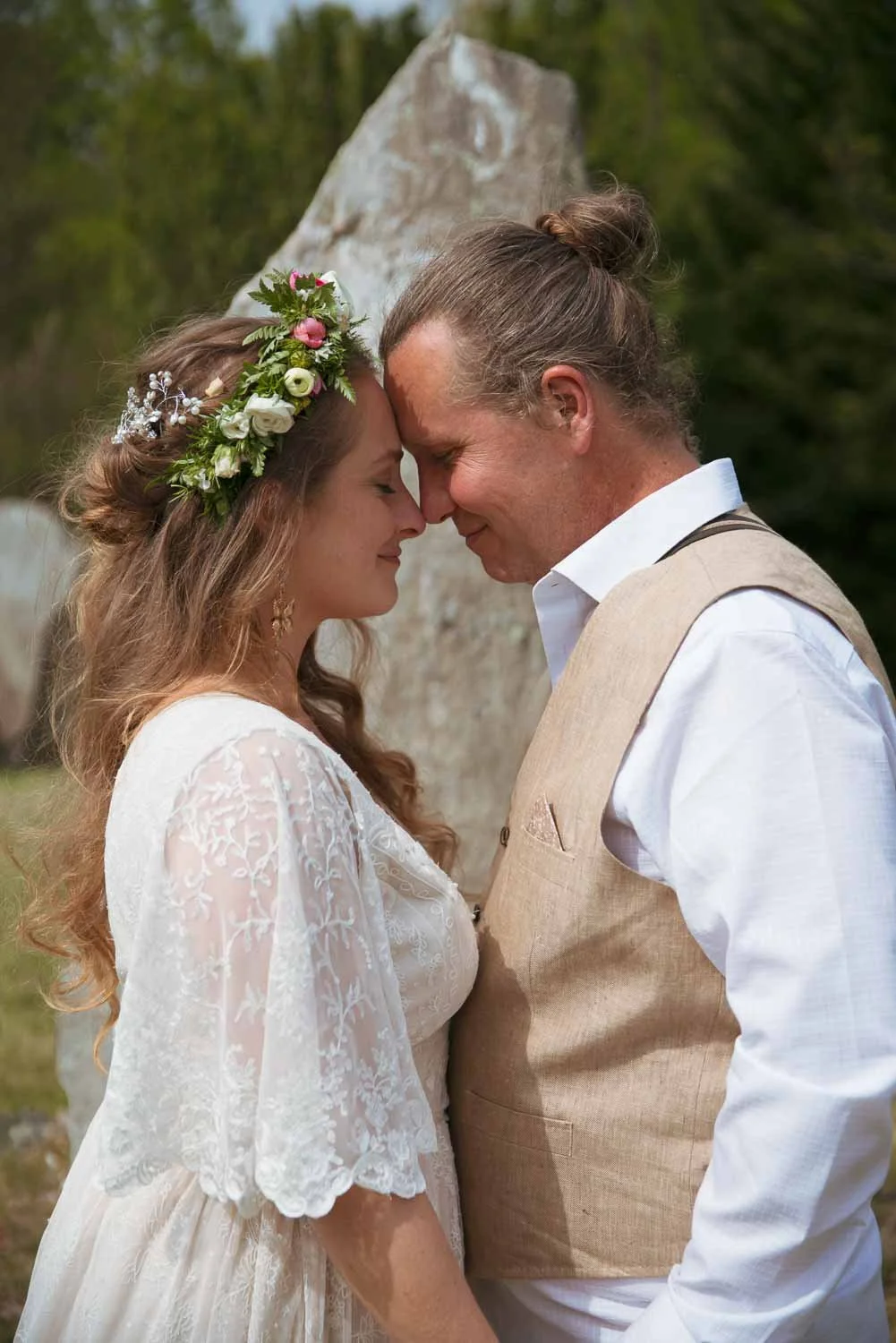A bride and groom with foreheads touching, eyes closed, outside during their wedding, with a large stone or rock in the background.