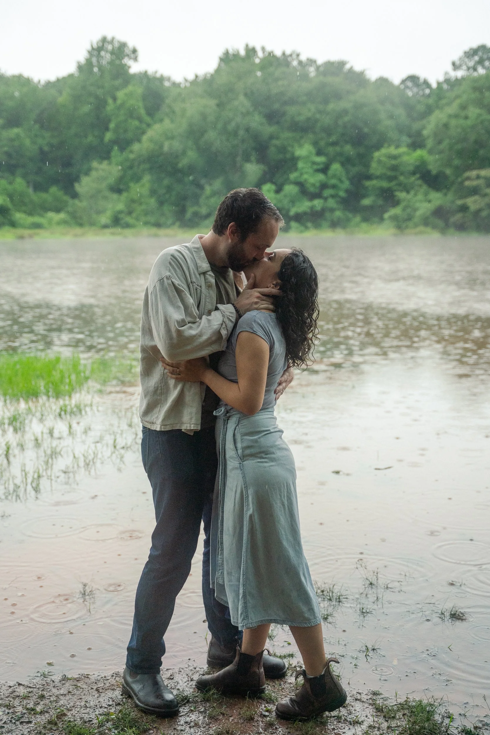 A couple sharing a kiss by a lake during rain, with lush green trees in the background.