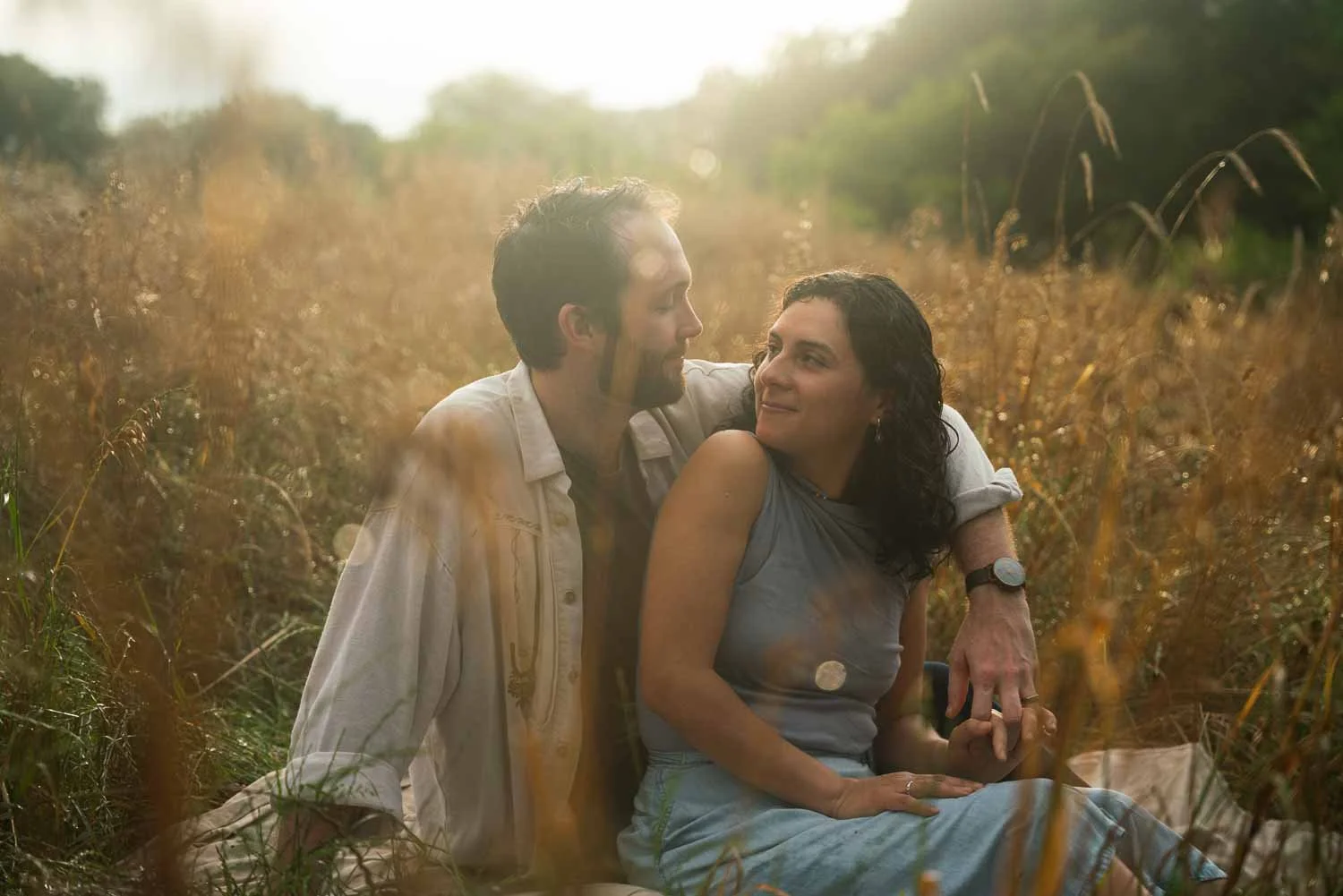A man and woman sitting closely together in a field of tall grass during sunset, gazing lovingly at each other.