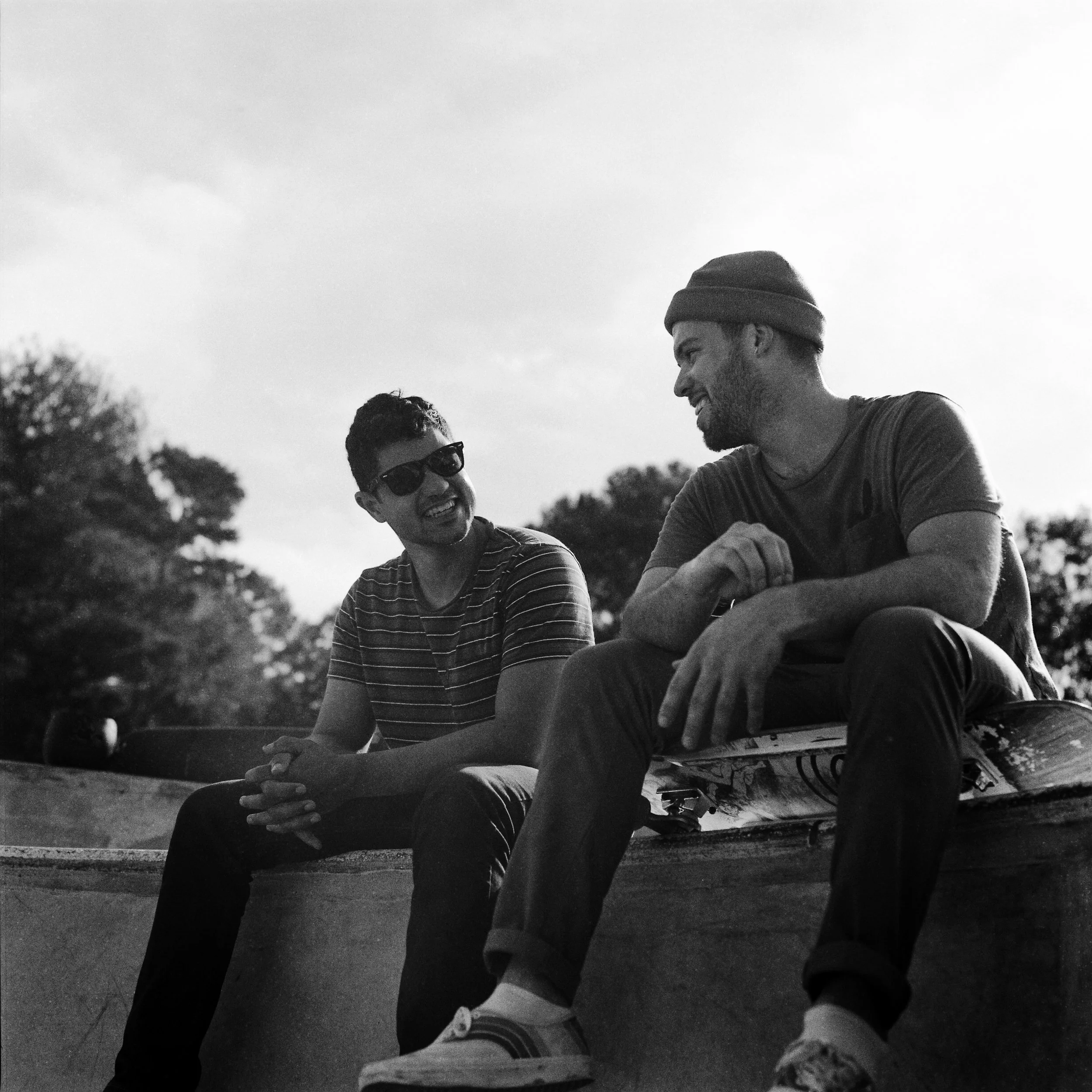 Two young men sit together at a skate park, smiling and talking, with one wearing sunglasses and the other wearing a beanie, in black and white photograph.