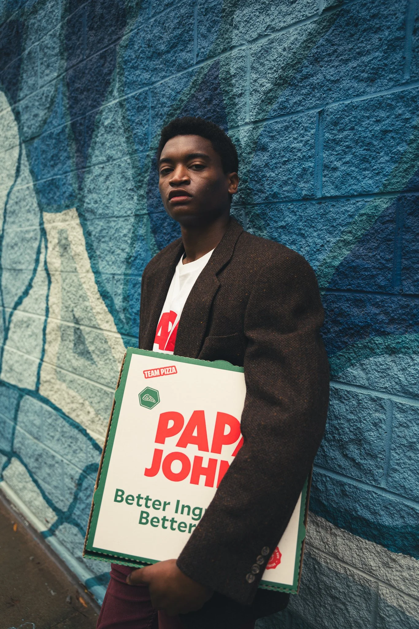A young man with dark skin and short curly hair standing against a blue graffiti wall, holding a pizza box with the words "Papa John's Better Ingredients Better Pizza" printed on it.