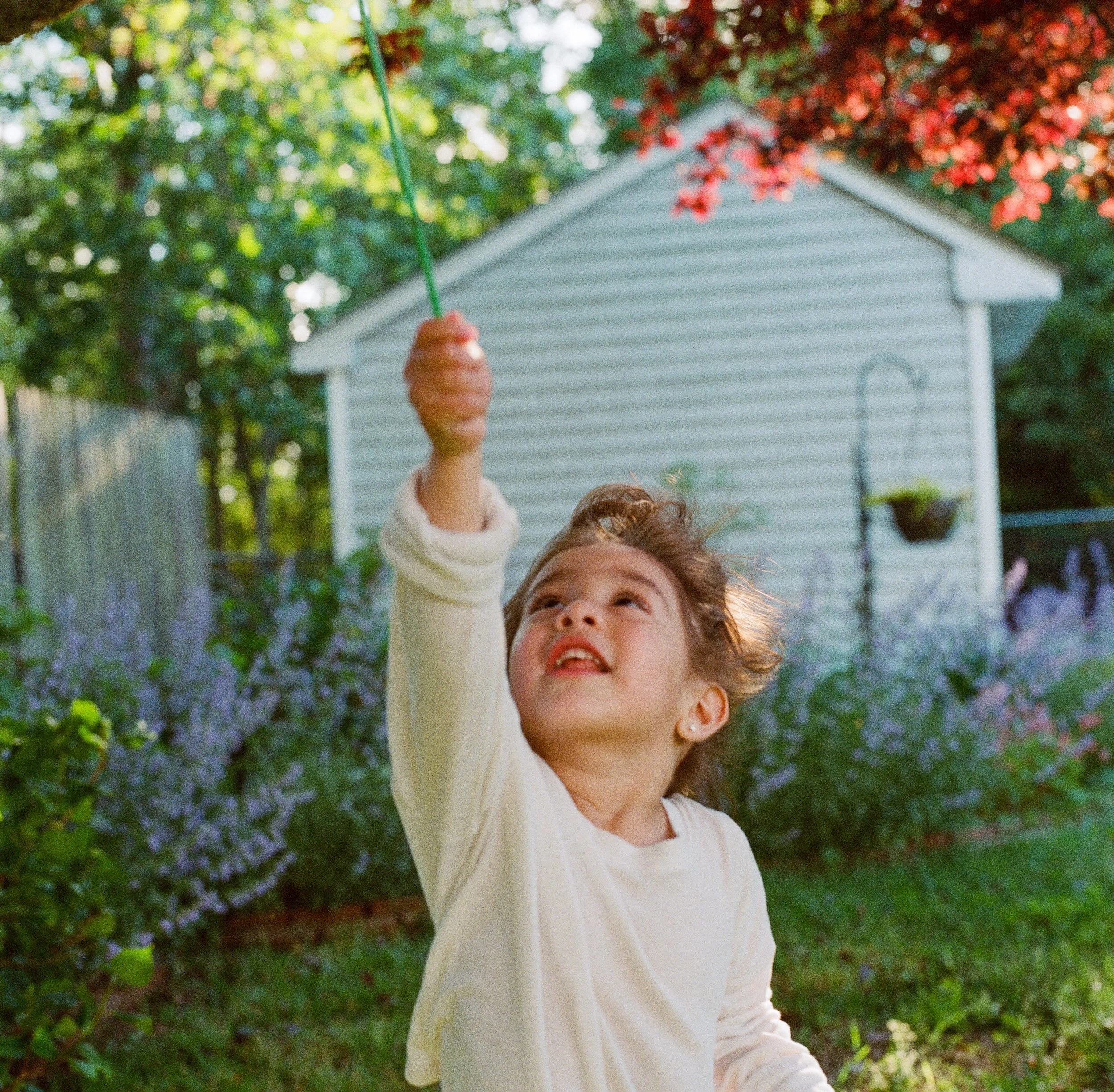 A young girl with curly hair playing with a bubble wand outdoors in a garden, with a white house and colorful bushes in the background.