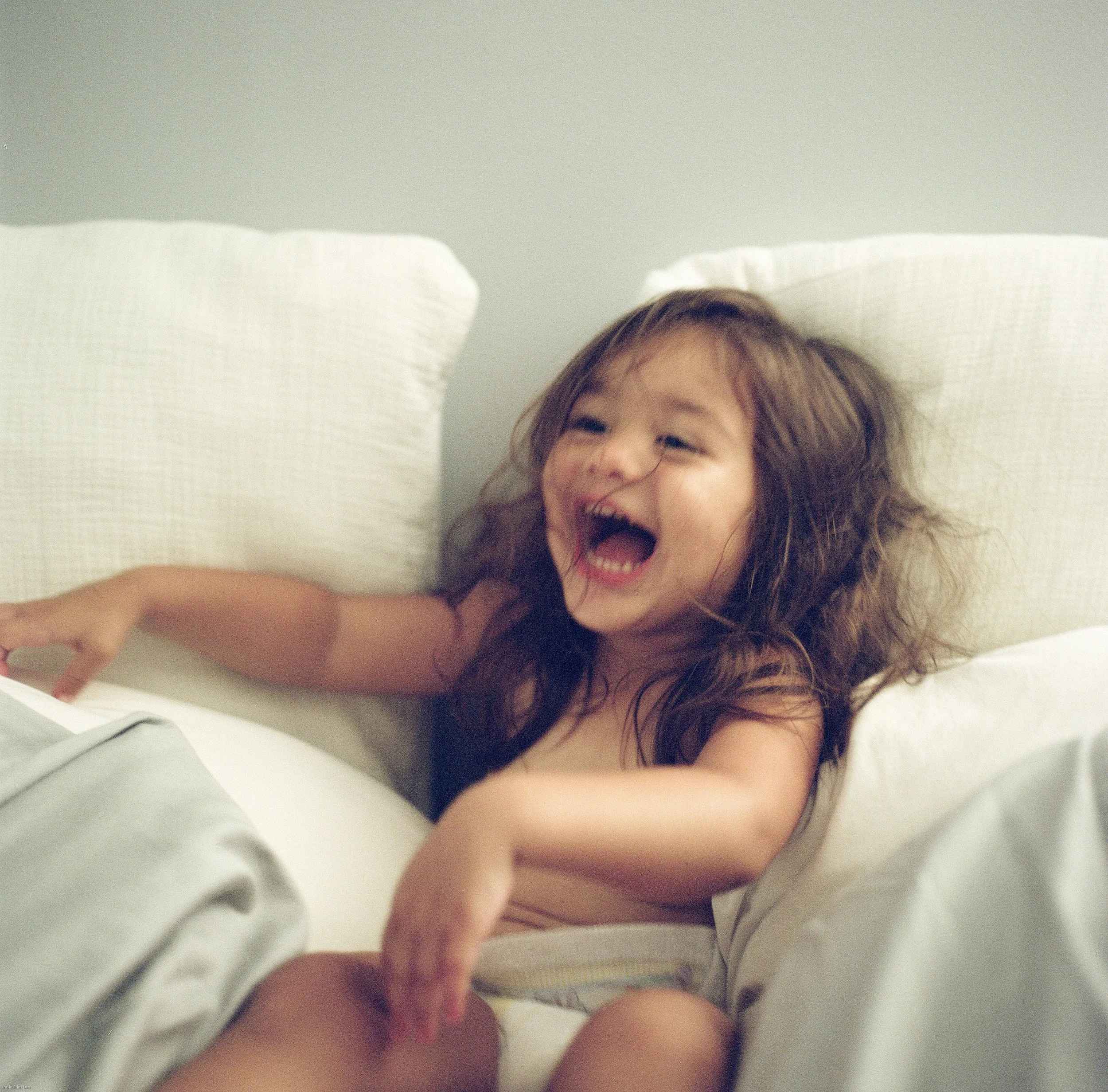 A young girl with long, curly hair, laughing while lying in bed with white pillows and sheets.