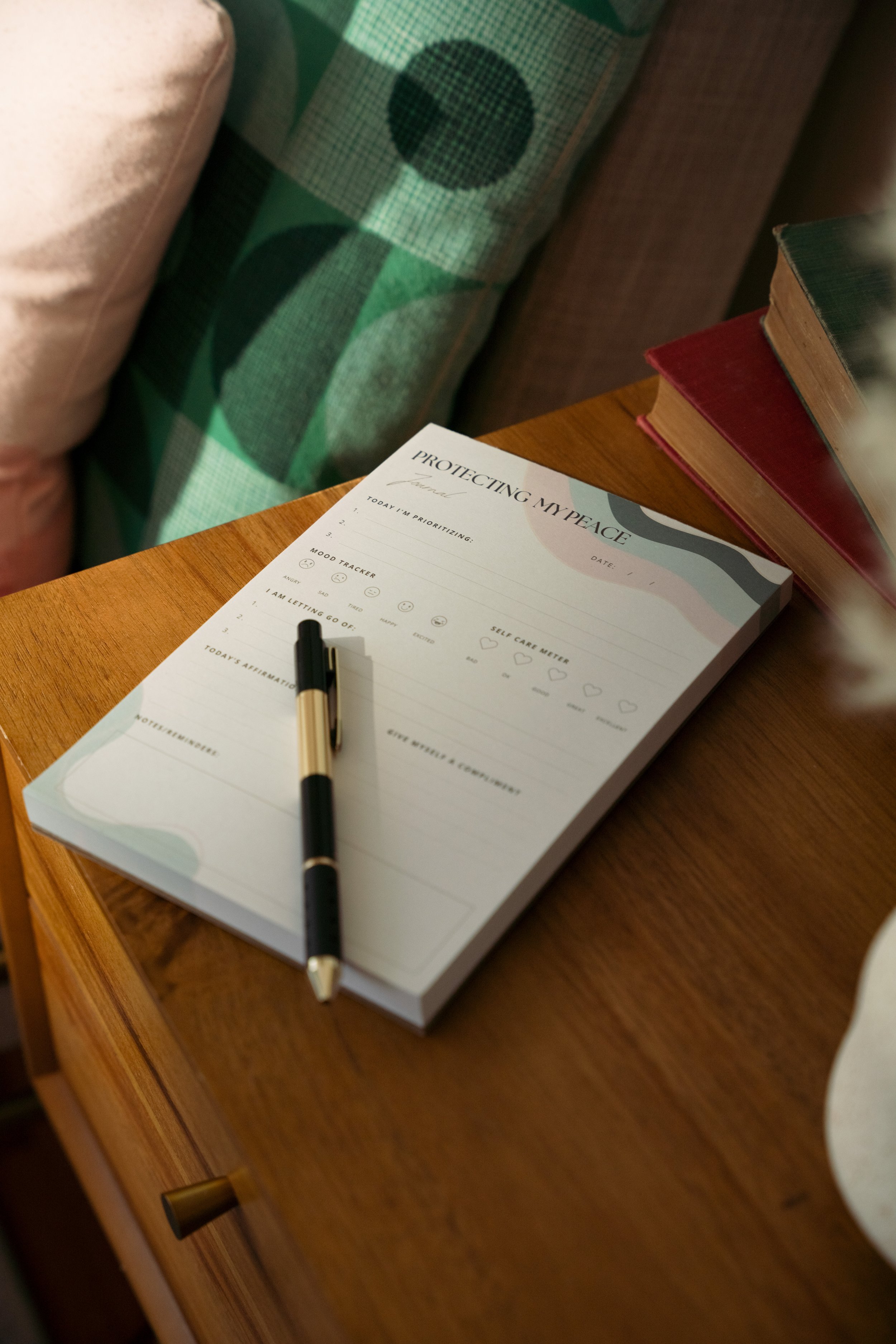 A to-do list and self-care tracker on a wooden nightstand, with a black and gold pen resting on it. The list is titled 'Protecting My Peace' and has sections for prioritizing, mood tracker, self-care meter, affirmations, and a quote.