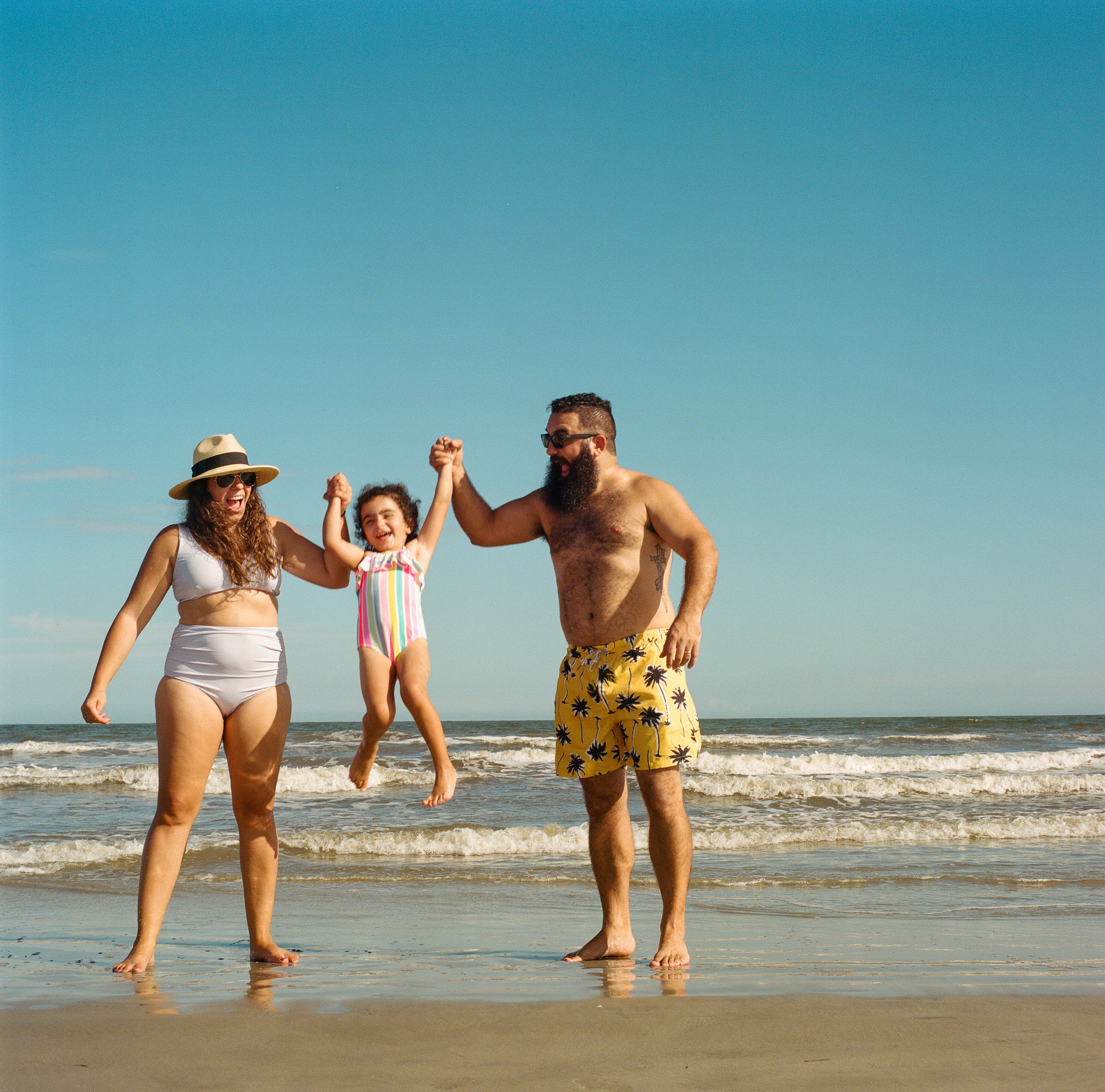 A family of three enjoying a day at the beach. The woman and man are holding a young girl between them, lifting her up and smiling. The woman is wearing a white bikini and a wide-brimmed hat, while the man is shirtless, wearing yellow shorts with a p