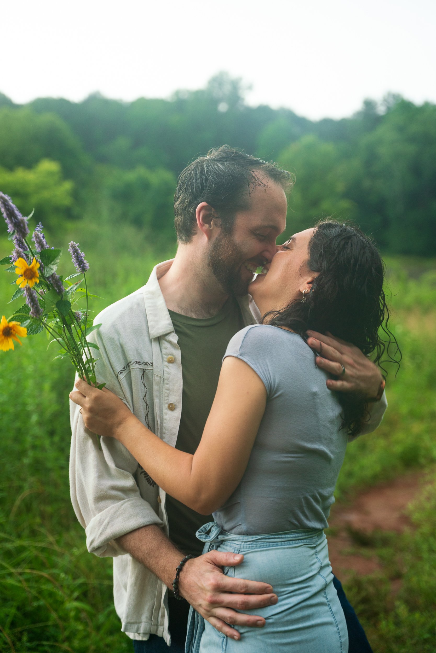 A couple sharing an embrace outdoors, the man holding a bouquet of colorful flowers, with a green forested background.