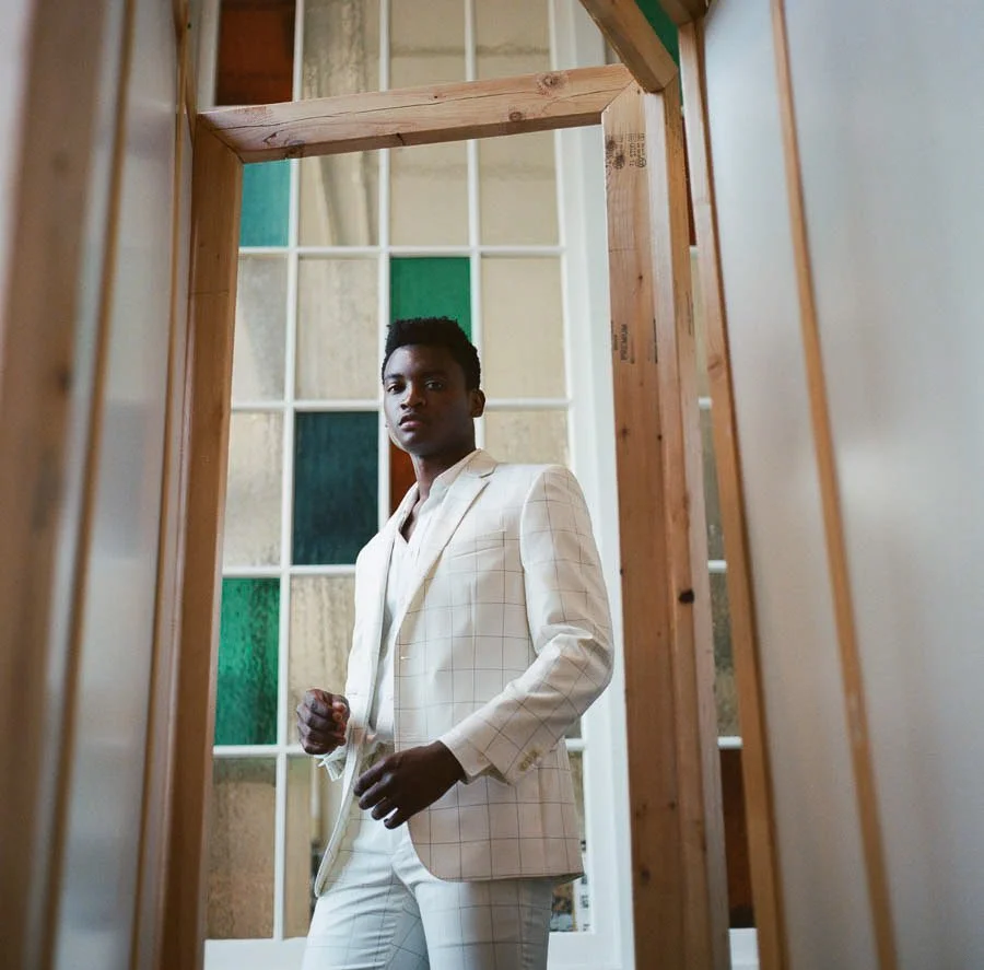 A man in a white suit with a checkered pattern stands in front of a stained glass window, looking at the camera, framed by a wooden structure.