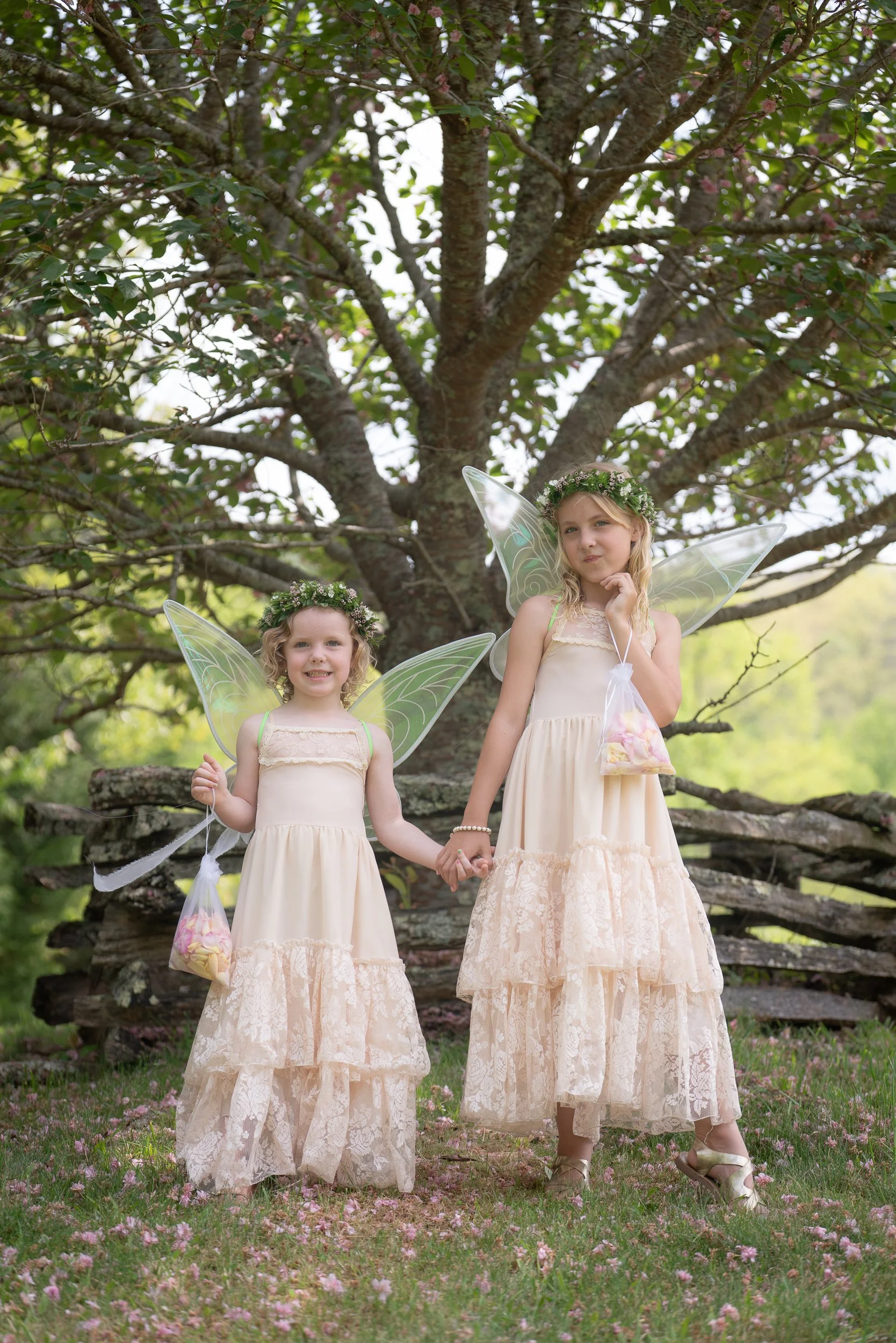 Two young girls dressed as fairies with wings holding hands outdoors under a large tree, wearing flower crowns and cream-colored dresses with lace, each holding a bag of candy.