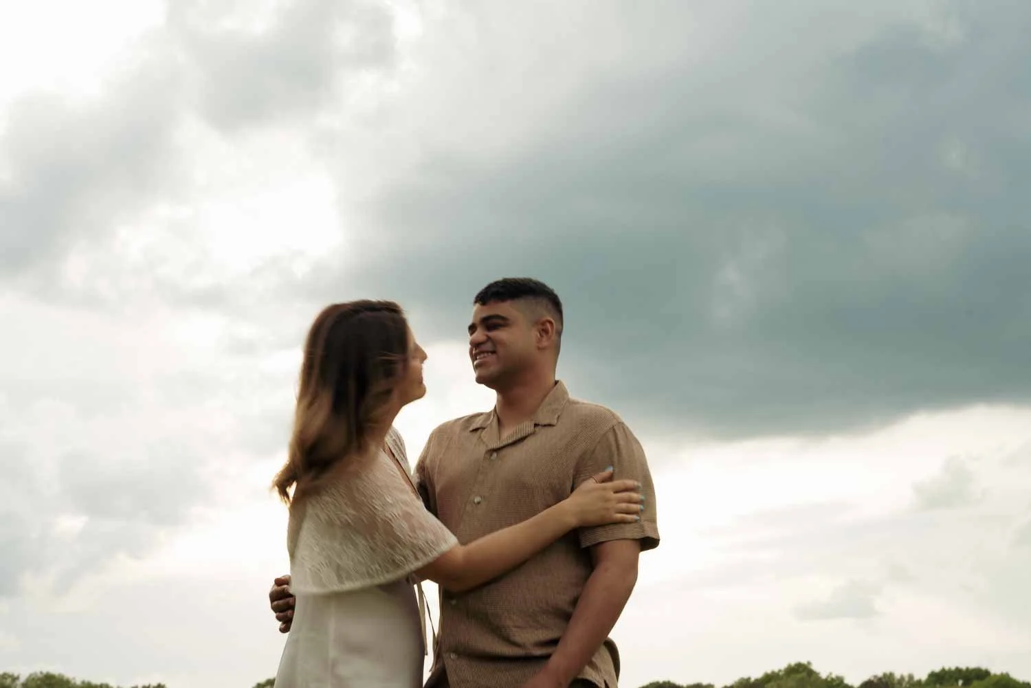 A couple embracing outdoors under cloudy sky, smiling at each other.