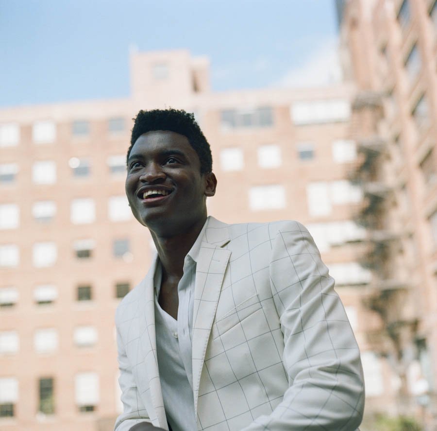 A young man smiling outdoors in front of tall apartment buildings, dressed in a light-colored checkered blazer and white shirt.
