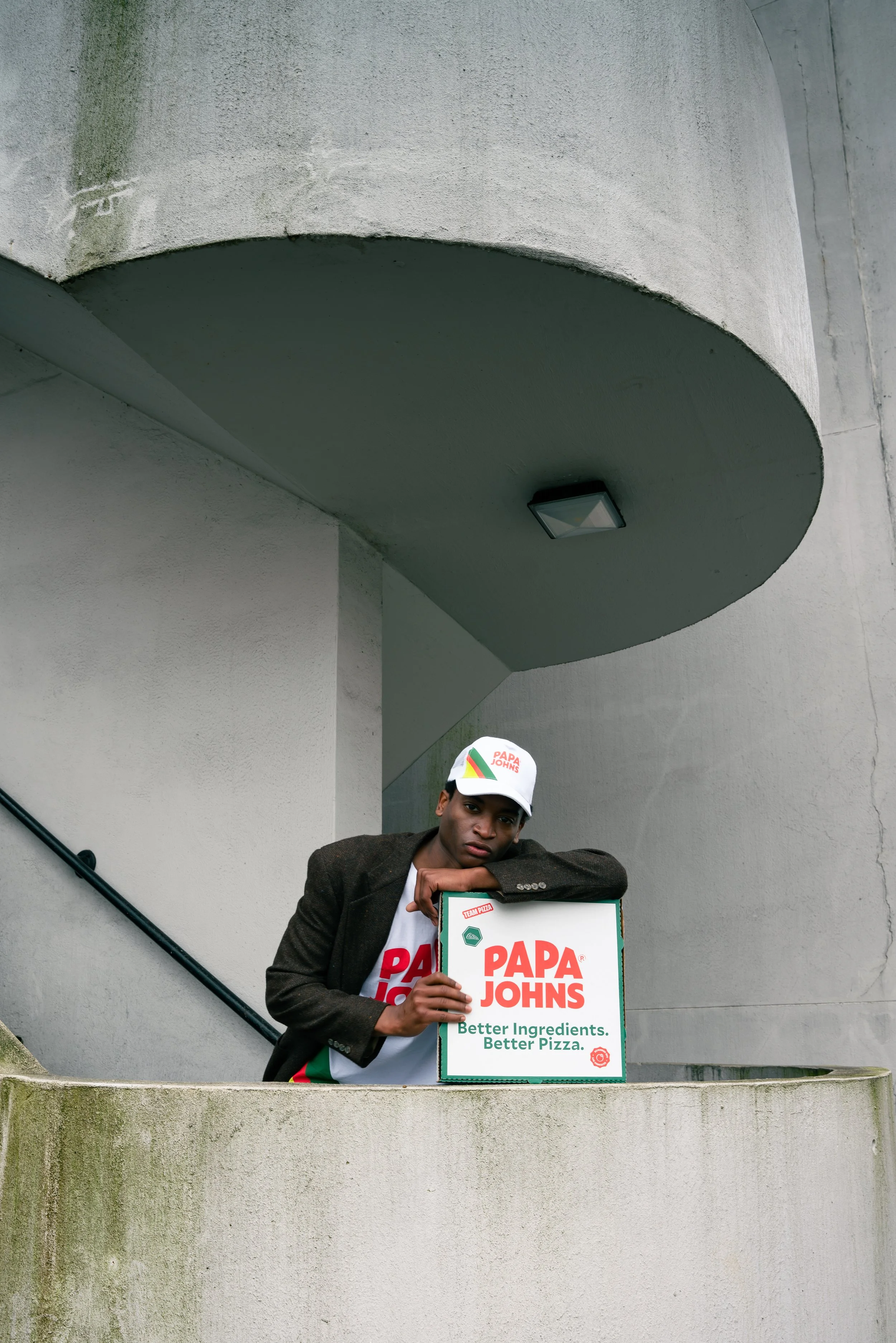 A young man leaning on a concrete ledge, holding a Pizza box from Papa John's, beneath an overhang of a modern building.