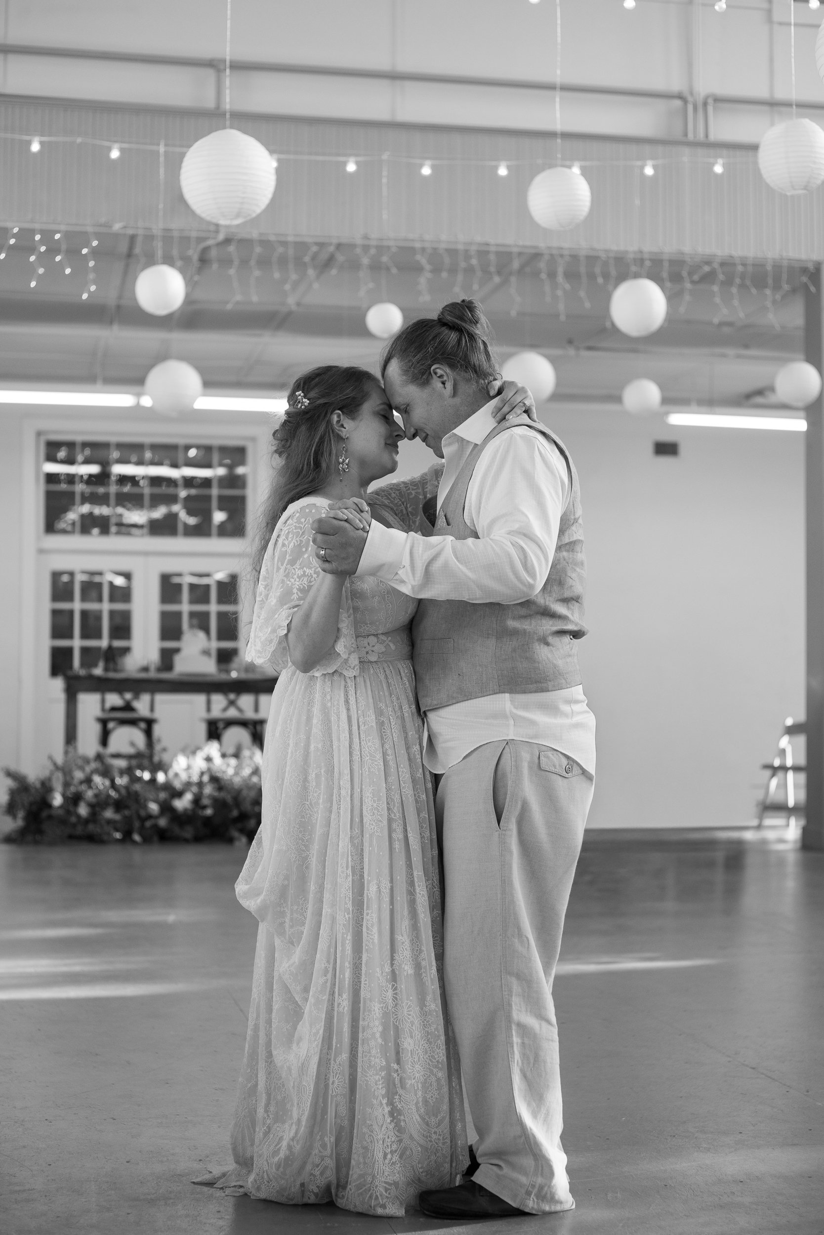 A couple is dancing closely at their wedding reception, embracing and touching foreheads, in a decorated indoor venue with lanterns and string lights.