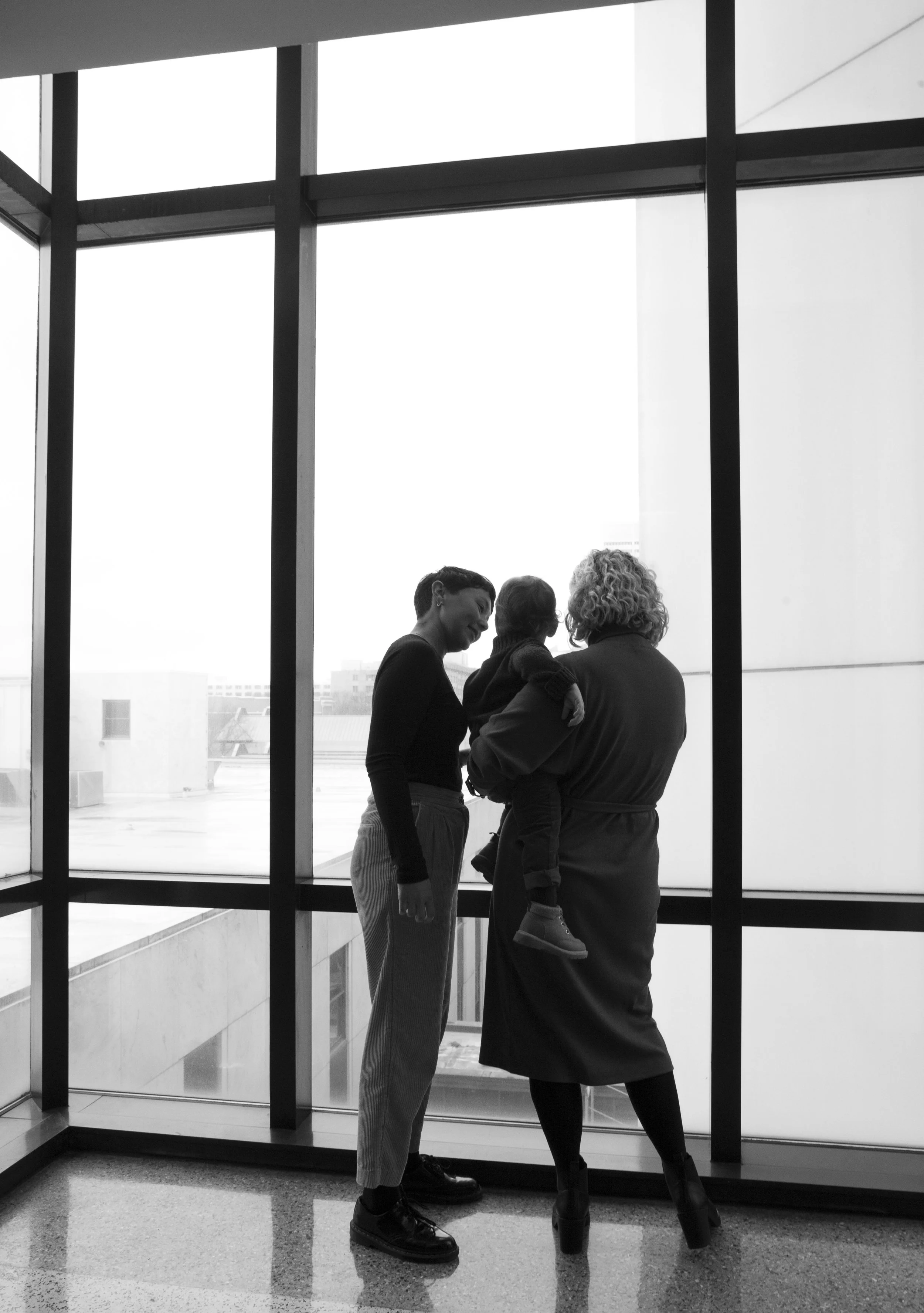Three people, two adults and a child, standing on an indoor balcony near large windows with a cityscape view. One adult holds the child, and all are engaged in conversation.