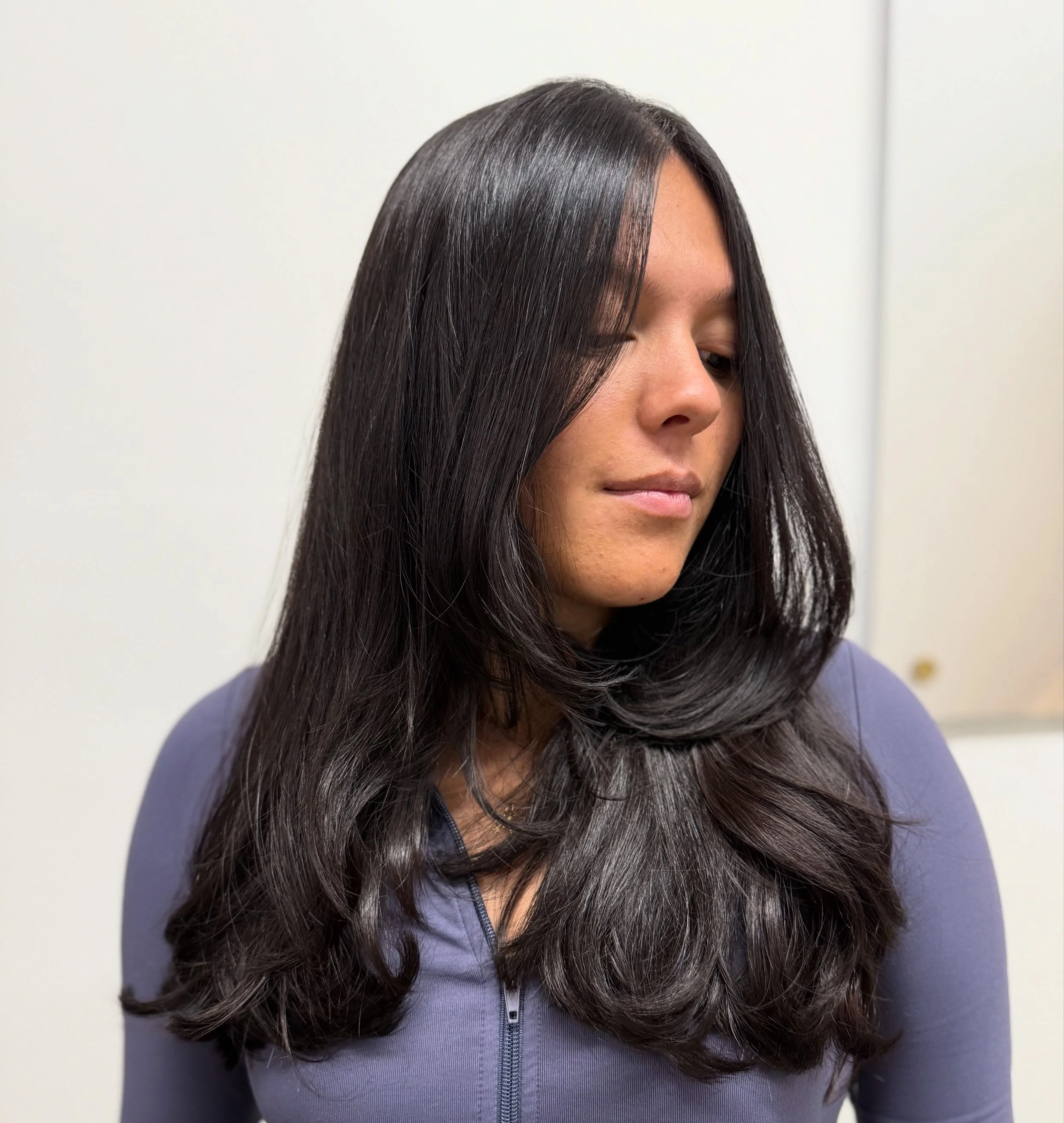 A woman with long, dark, shiny hair wearing a gray zip-up top, looking down with a neutral expression.