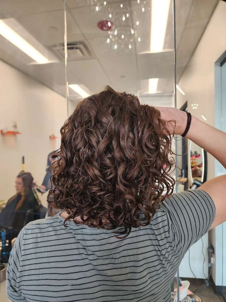Woman with shoulder-length, dark brown, curly hair in a salon, seen from the back, wearing a gray and black striped shirt.