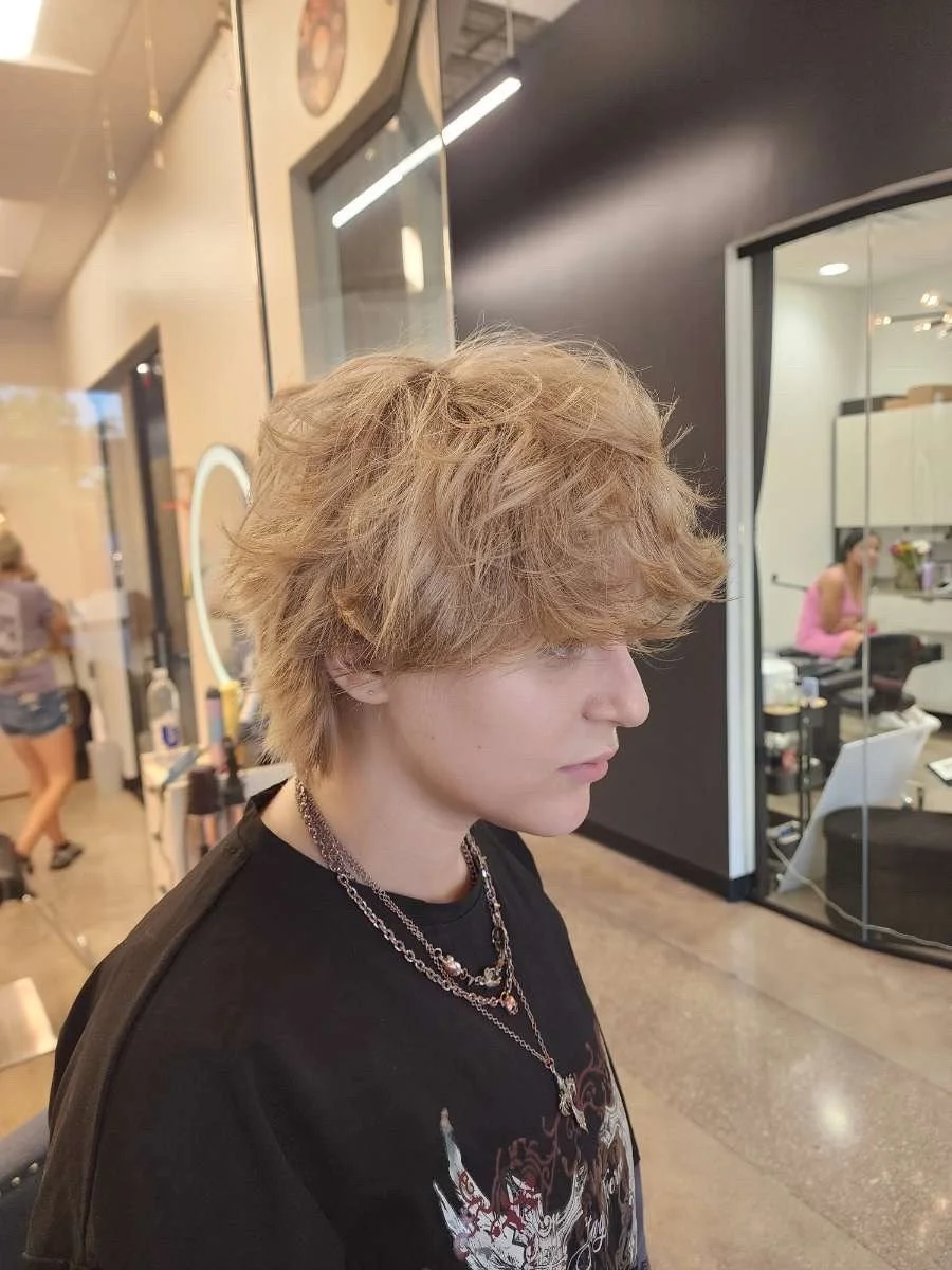 A young person with tousled blonde hair sitting inside a salon, wearing layered necklaces and a black graphic T-shirt. In the background, a salon worker and another customer are visible.