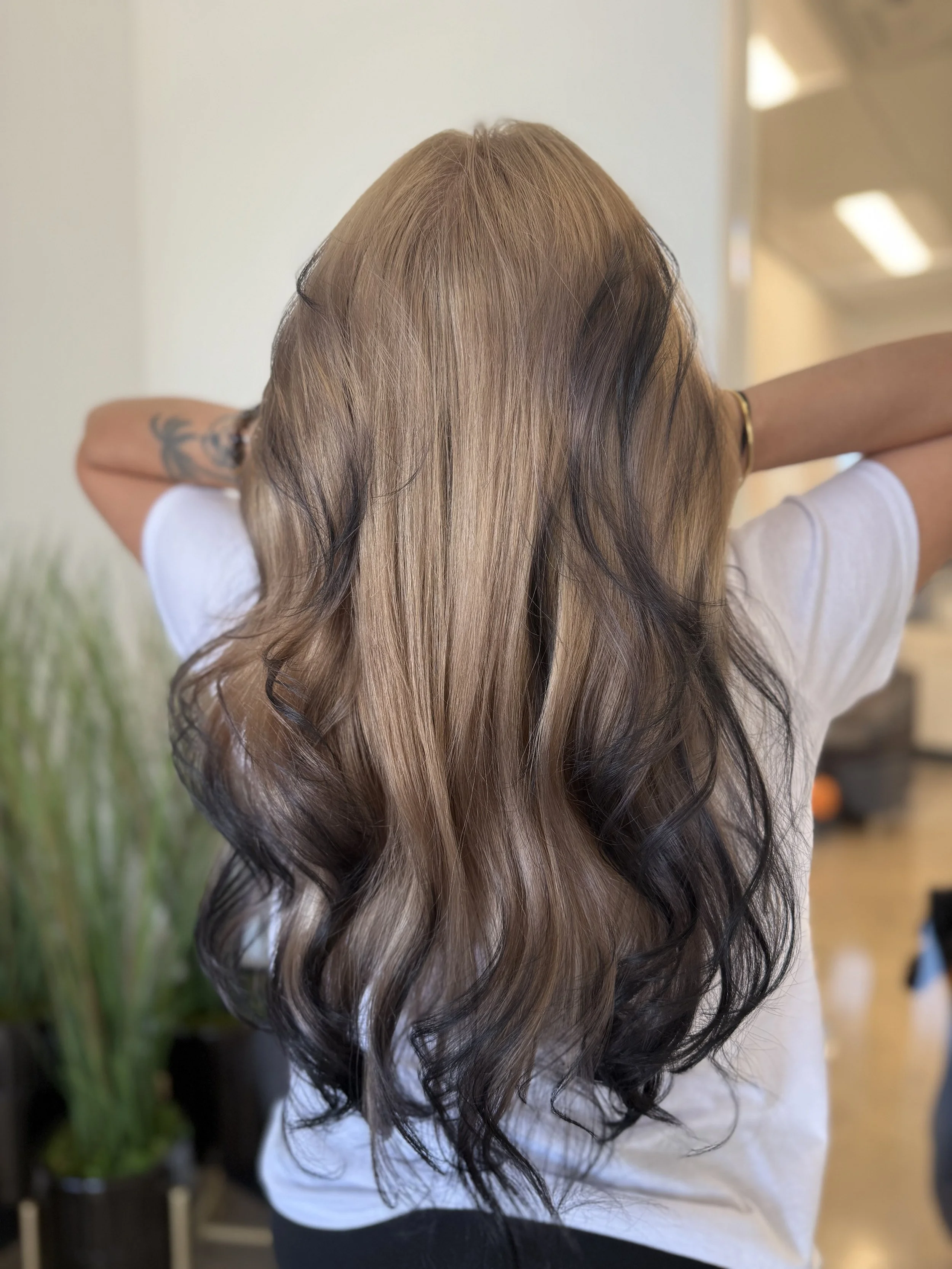 A woman with long, wavy hair featuring brown and black highlights, seen from the back with arms raised behind her head.