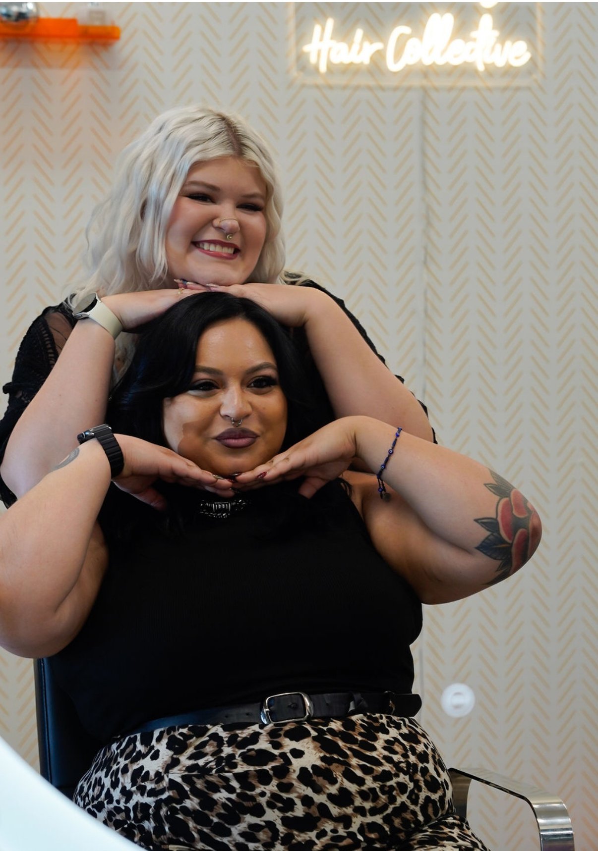 Two women posing together at a hair salon, with a sign in the background that reads 'Hair Collective.' The woman in front has dark hair, tattoos on her arm, and is wearing a black top and leopard print bottom. The woman behind has platinum blonde hair and is smiling cheerfully.