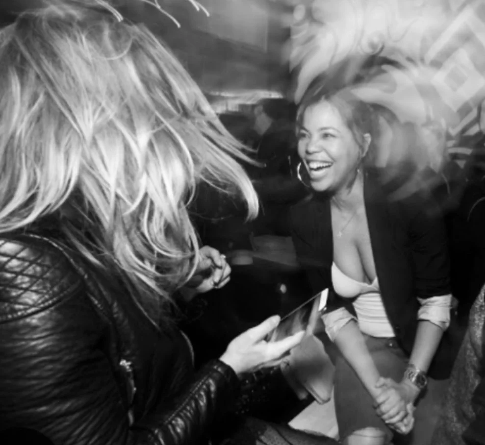 Two women smiling and laughing while sitting indoors. One woman with blonde hair is wearing a leather jacket and holding a smartphone, the other woman with dark hair is wearing a blazer over a top with a deep neckline and is holding her hands together.