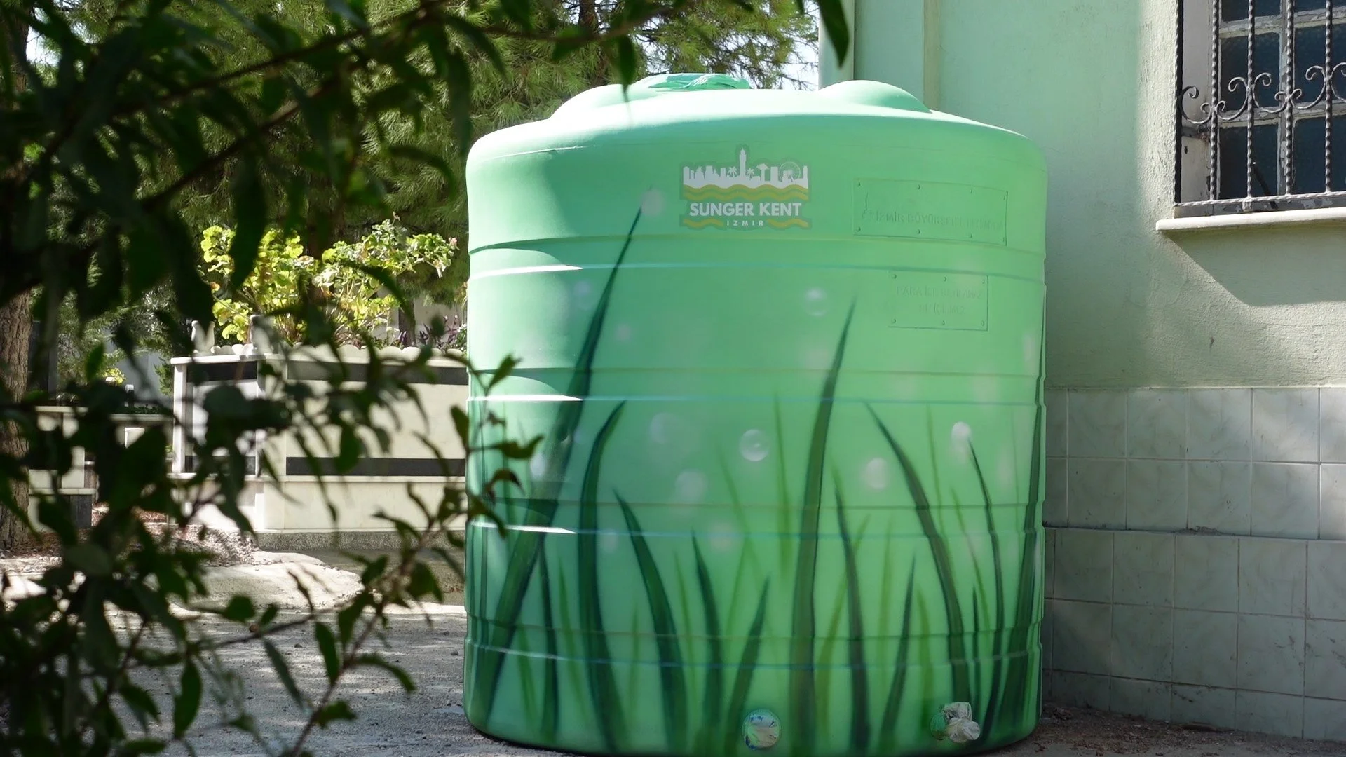 Yeşil su tankı dış mekanda, üzerine çim ve gökyüzü resmiyle süslenmiş, yakınındaki duvar ve çiçekli bitki.