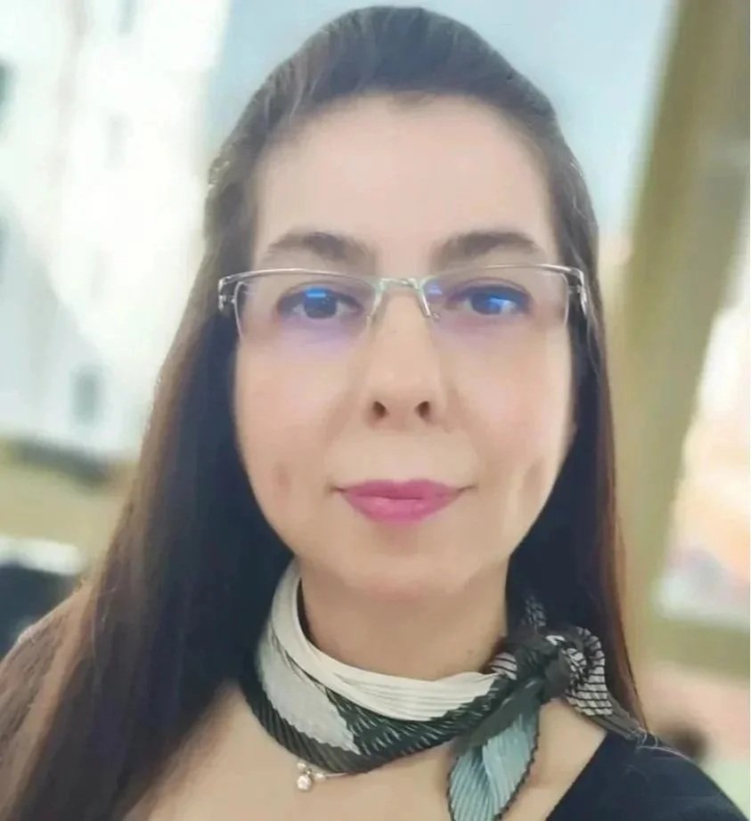 Kadın, renkli saç fuları ve gözlük takmış, ofis ortamında gülümseyerek poz veriyor.