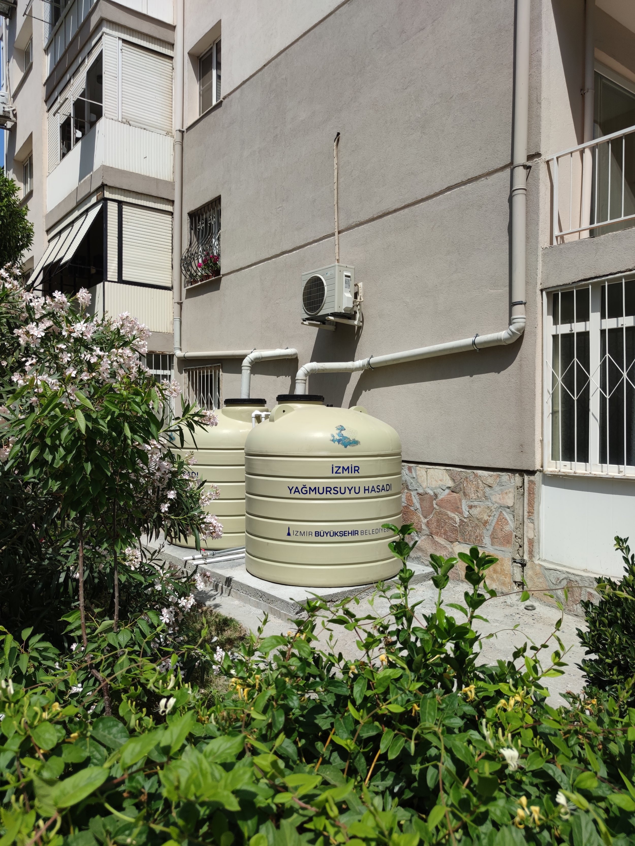 İki büyük su deposu, apartmanın dışında yer alıyor ve üzerinde İzmir Büyükşehir Belediyesi yazısı bulunuyor. Çevresinde çiçekler ve yeşil bitkiler var. Binanın duvarında klimalar ve pencereler yer alıyor.