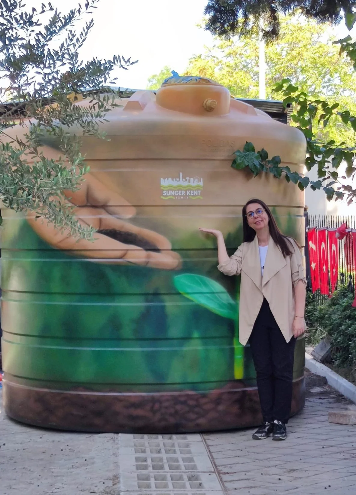 Dökme su tankının üzerinde bir kadın ve bazı ağaçlar ve bitkiler görünmekte. Tankta ve arka planda Türk bayrağı var.