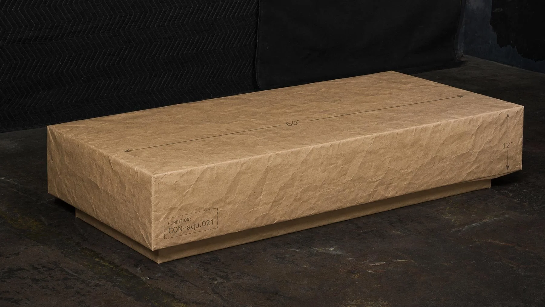 Paper-wrapped concrete coffee table with dimensional markings documenting condition CON-aqu.021 and a 60-inch monolithic form. action:wrapped