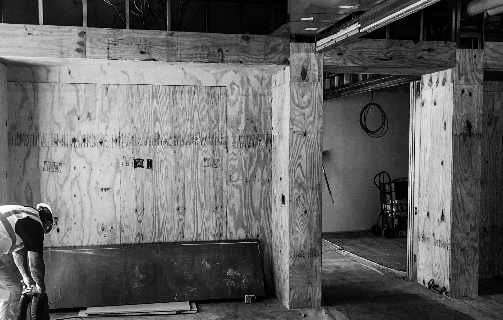 Construction site with wooden framing and plywood walls, a worker in safety gear handling a tool, and construction materials and tools in the background.