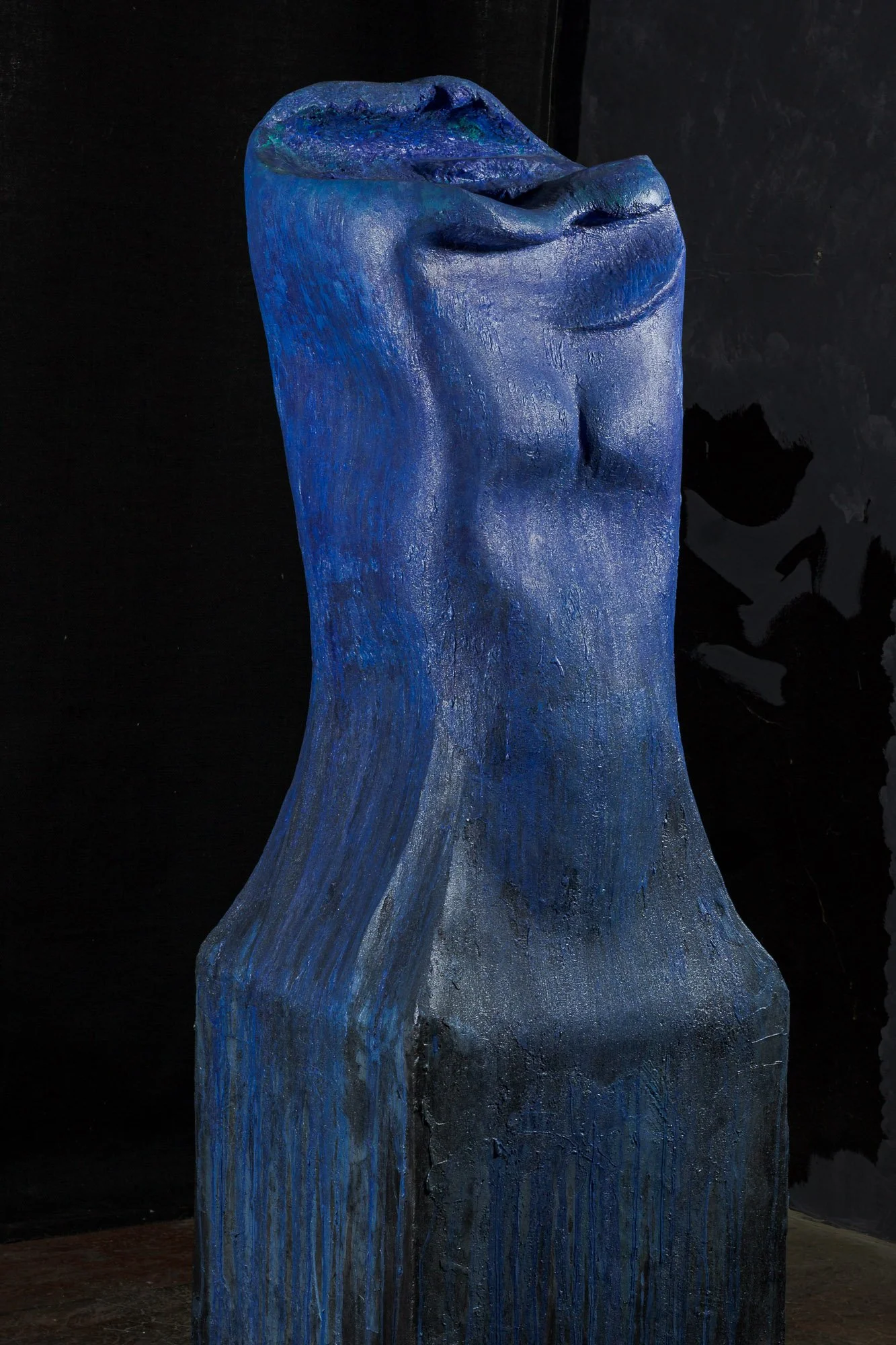 A blue, textured abstract sculpture resembling a distorted human face or mask with closed eyes, against a dark background.